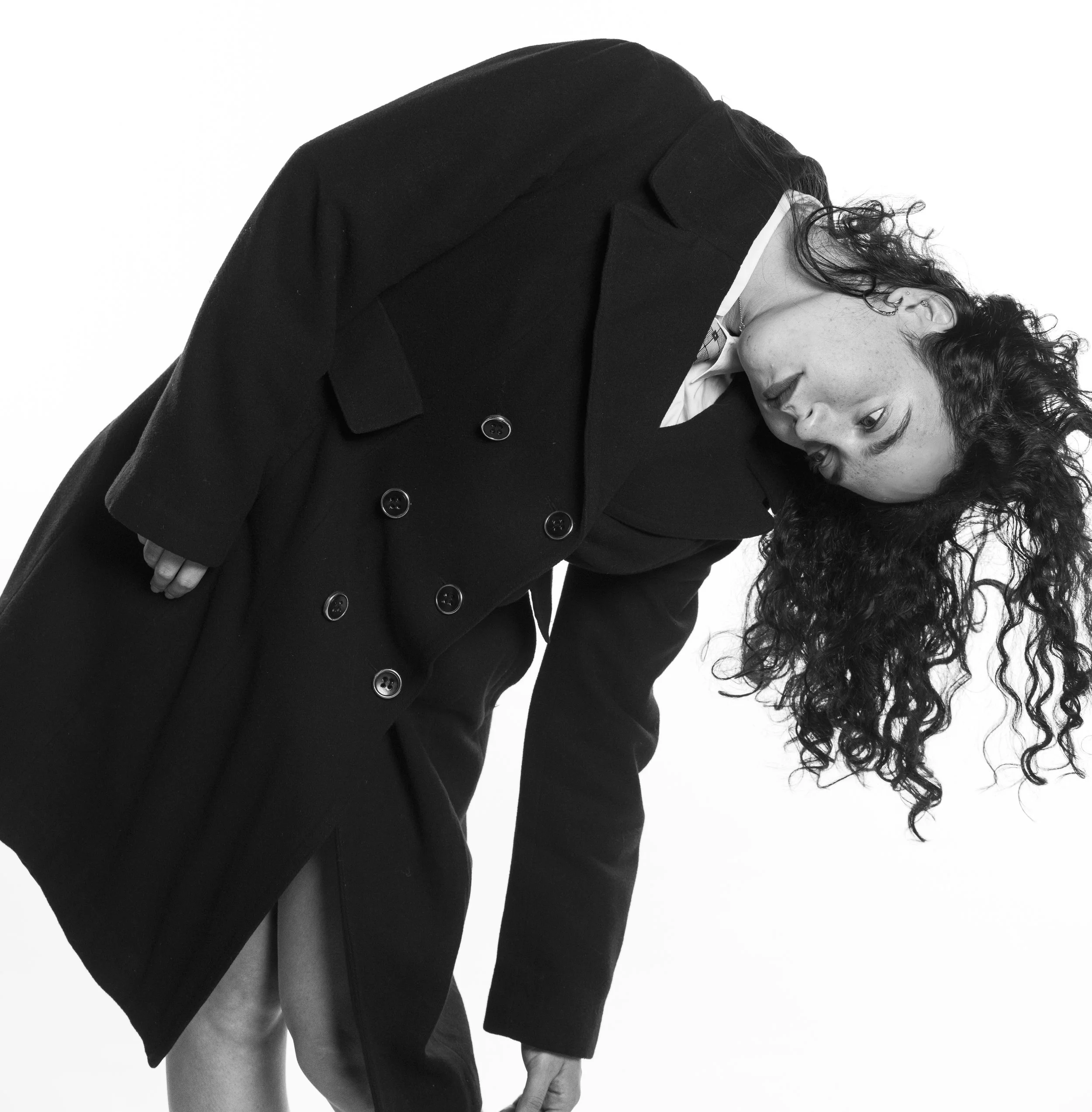 A woman with curly hair wearing a long black coat, bending forward with her head tilted to the side, against a plain white background.