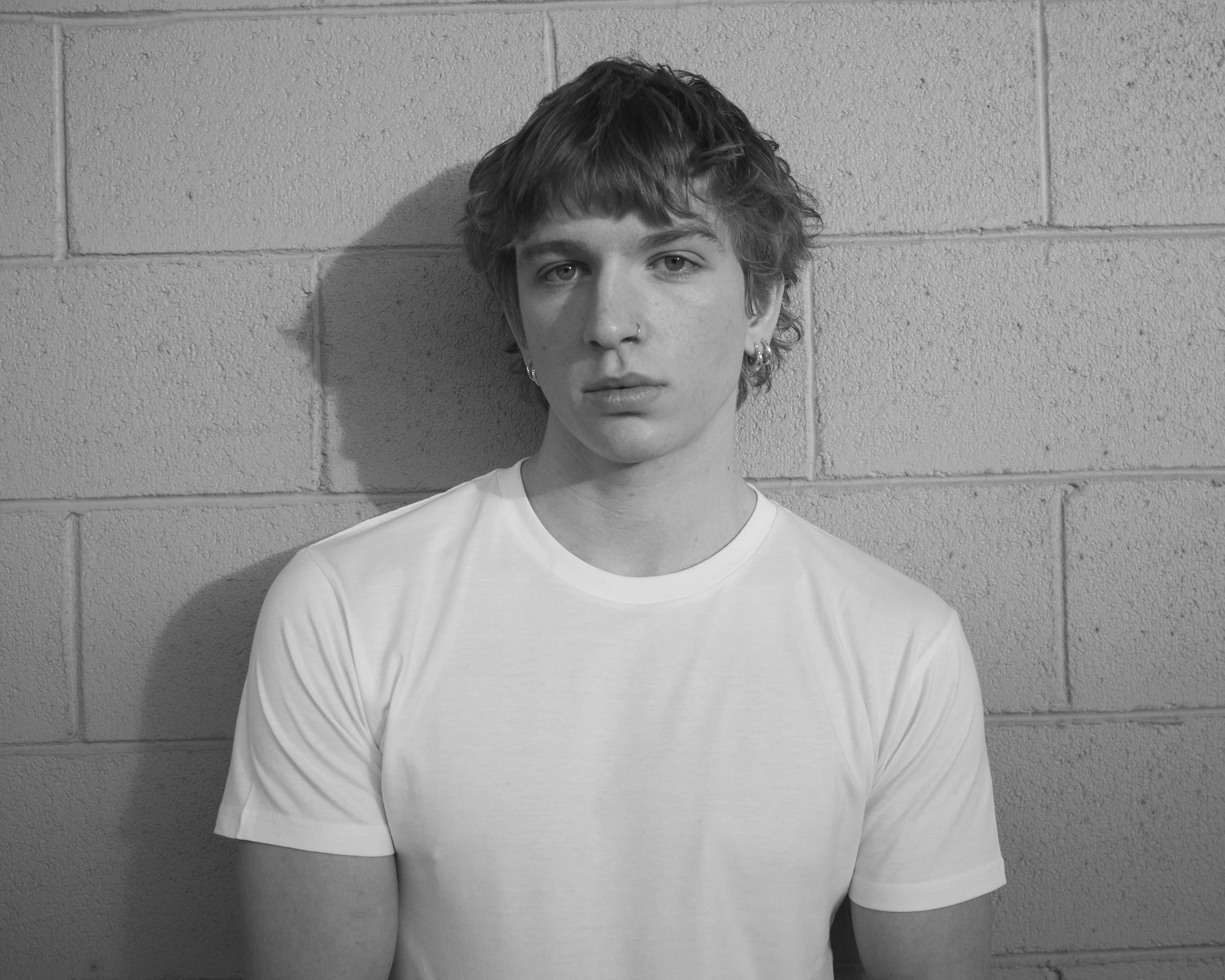 A young person with short hair, wearing a white t-shirt, standing against a brick wall with a shadow cast on the wall.