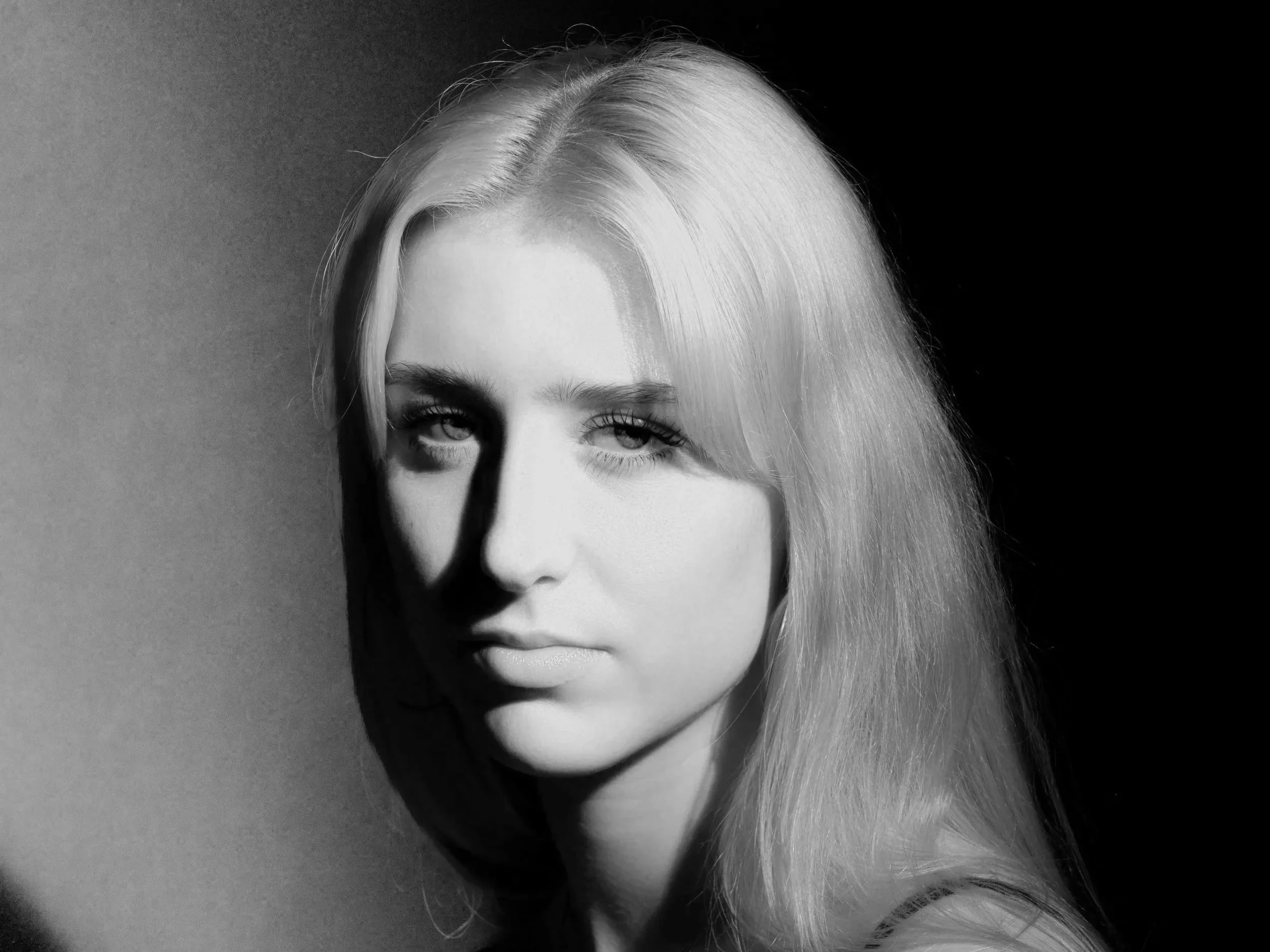 Black and white portrait of a woman with long blonde hair, looking at the camera with a serious expression, partially shadowed on one side.