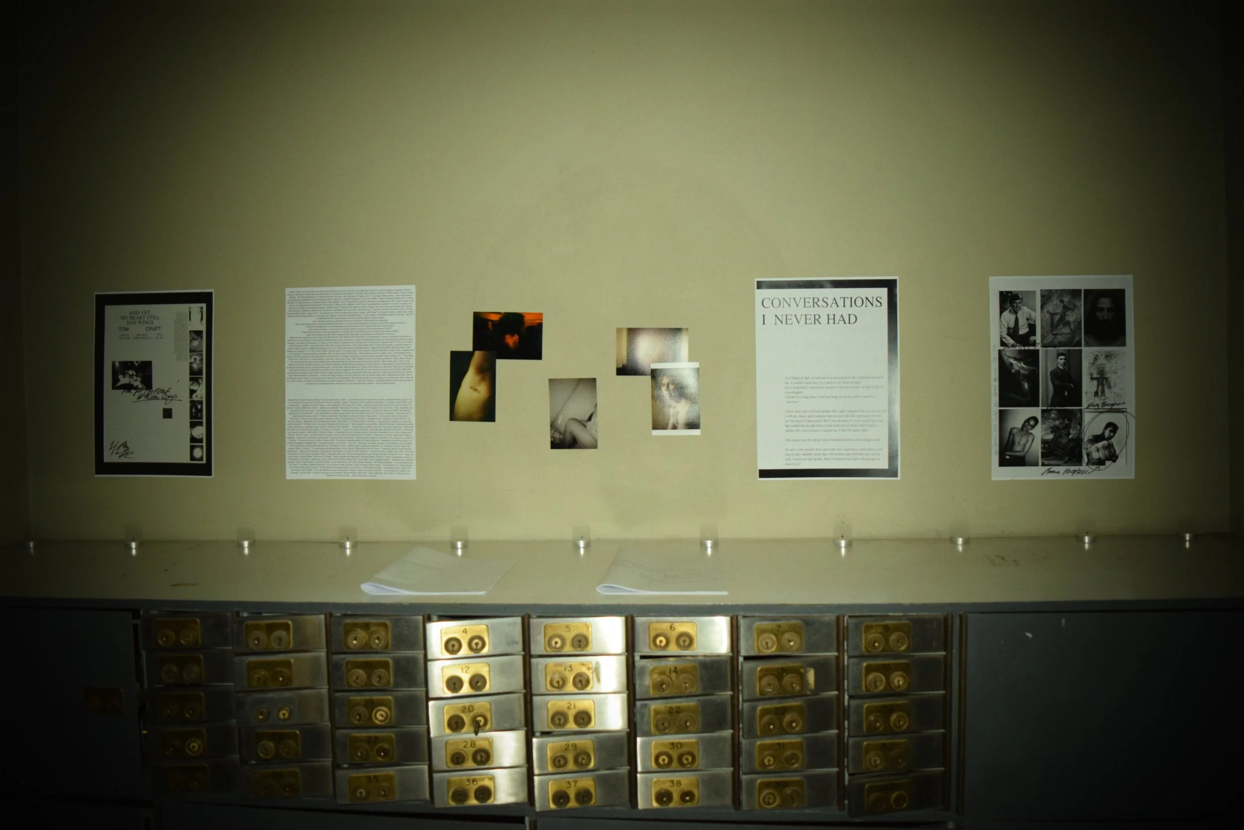 Display of photographs and text panels on a beige wall in an art exhibit, with a metal storage cabinet with numbered drawers below.