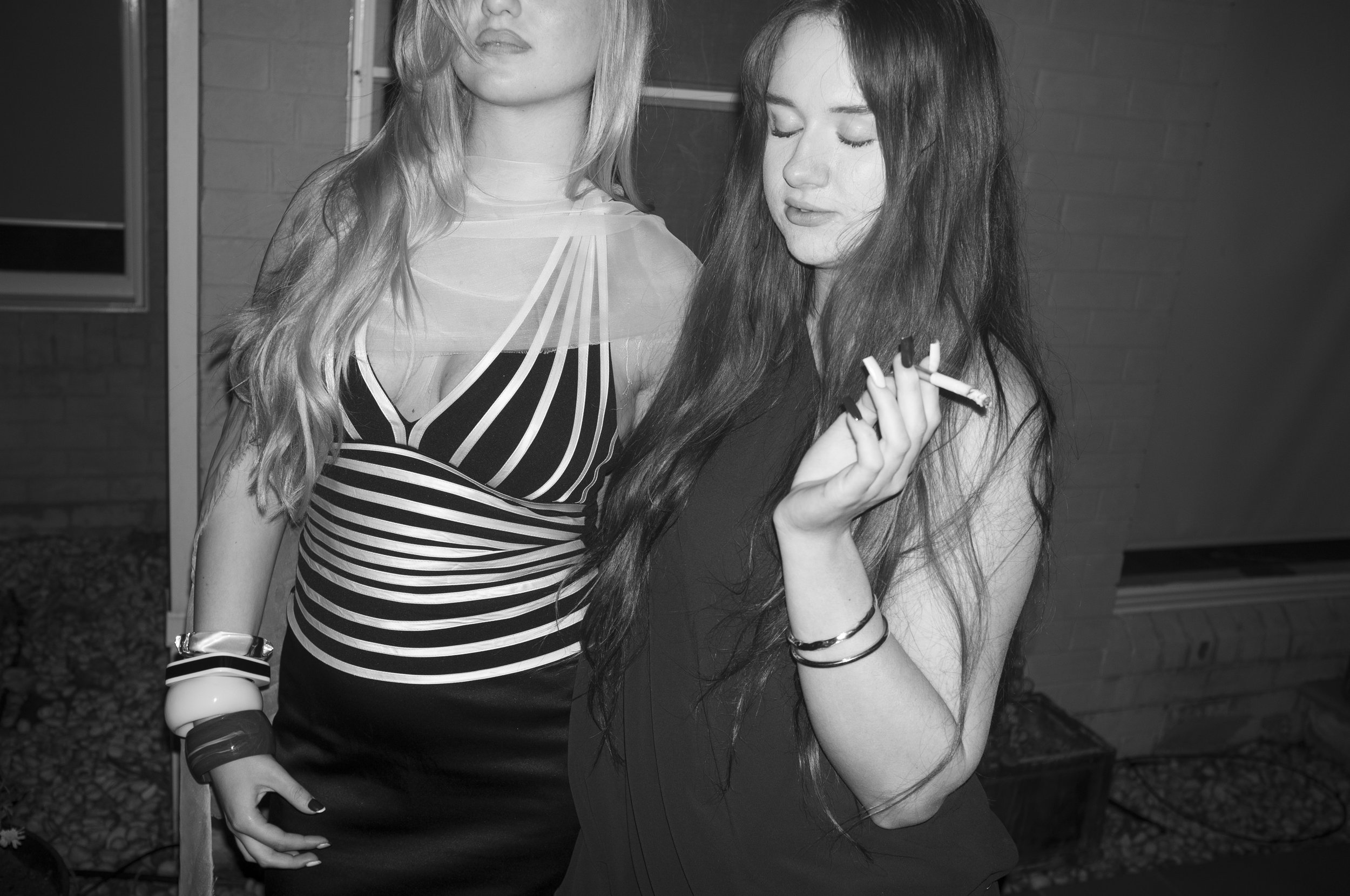 Two women pose together, one with light hair and the other with dark hair, both wearing stylish dresses. The woman with dark hair is holding a cigarette and has her eyes closed.