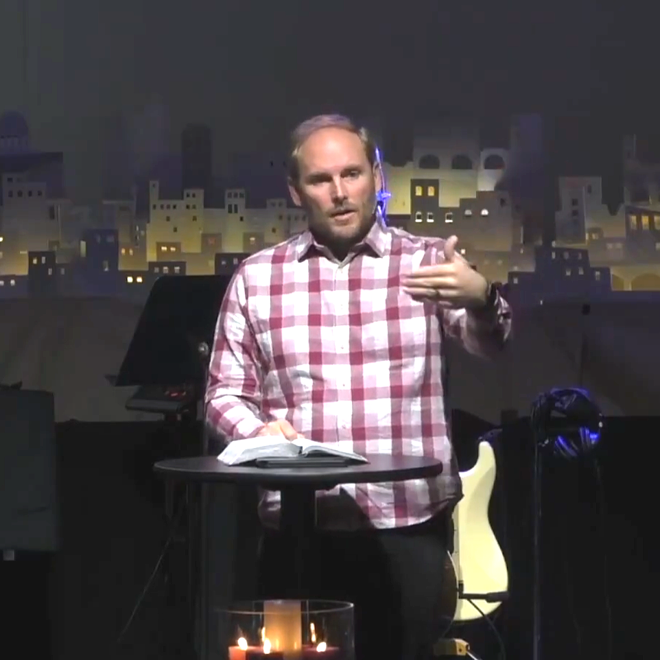 A man in a pink and white checkered shirt is giving a presentation or lecture, standing behind a small round table with an open book on it, in a setting with a cityscape backdrop and musical instruments nearby.