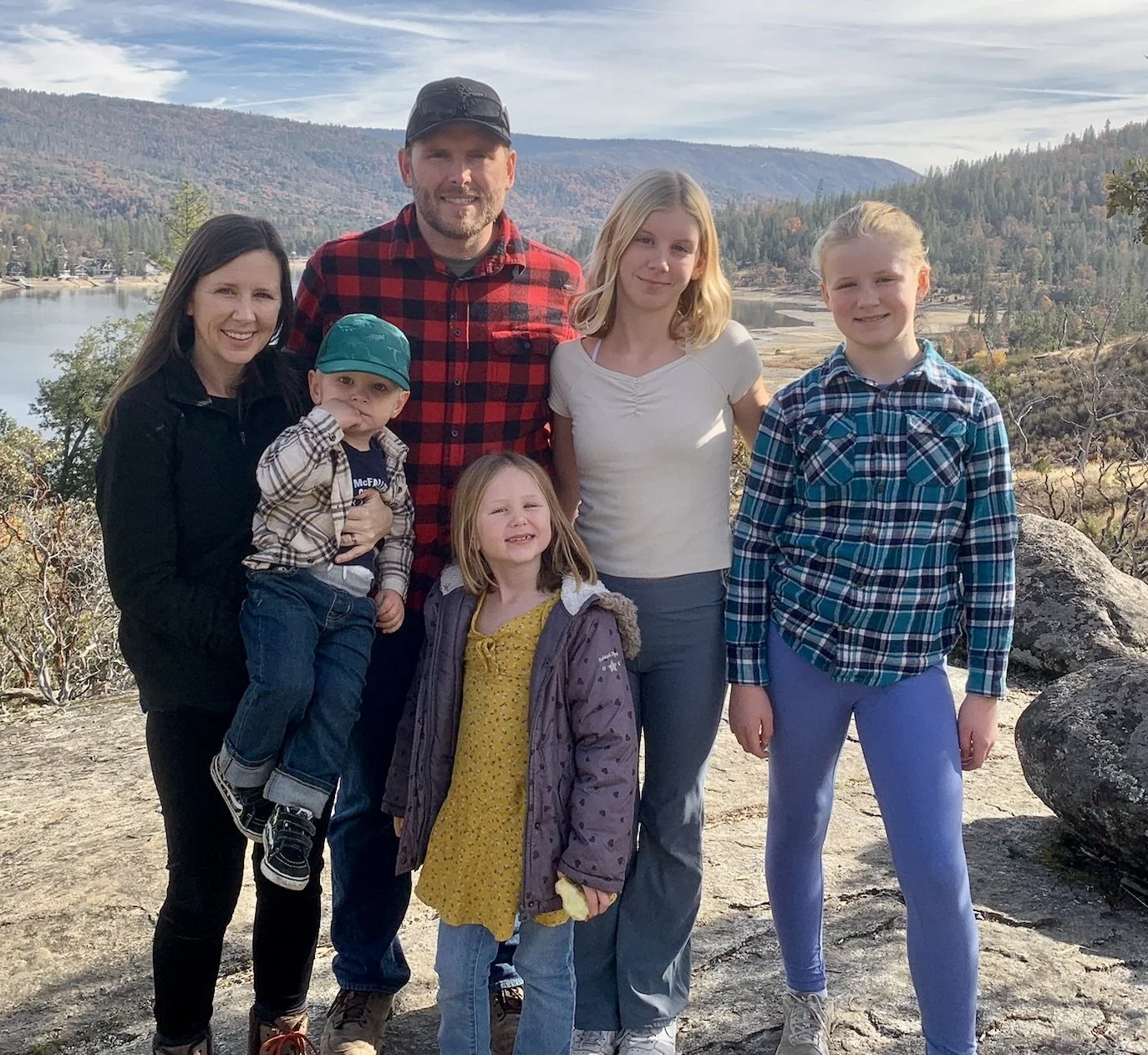 A family of seven standing outdoors on a rocky area with a lake and mountains in the background. The family includes two adult women, one adult man, and four children. They are dressed in casual clothing and are smiling at the camera.