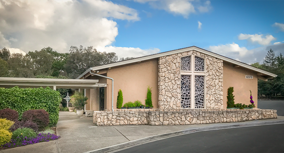 A church building with a large cross on the front window, surrounded by landscaped shrubs and flowers, under a partly cloudy sky.
