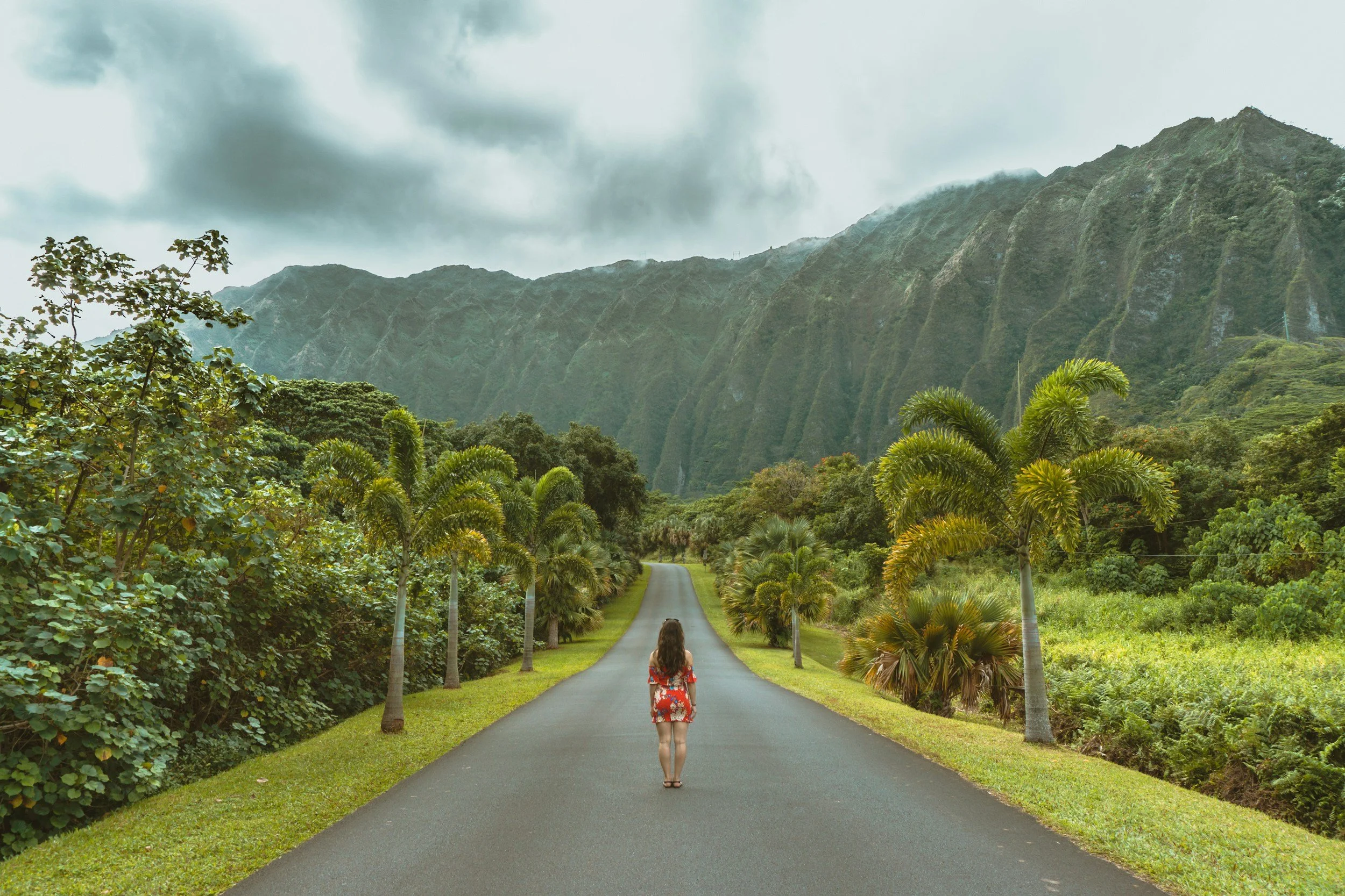 A woman in a red floral dress walking down an empty paved road surrounded by green palm trees and lush vegetation, with tall mountains in the background and overcast sky.