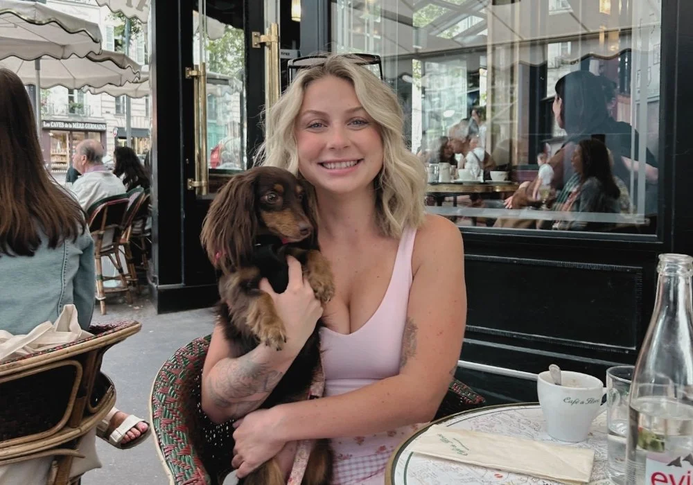 A woman with blonde hair smiling and holding a small brown and black dog in a cafe. The cafe has large windows and several other people sitting inside and outside.