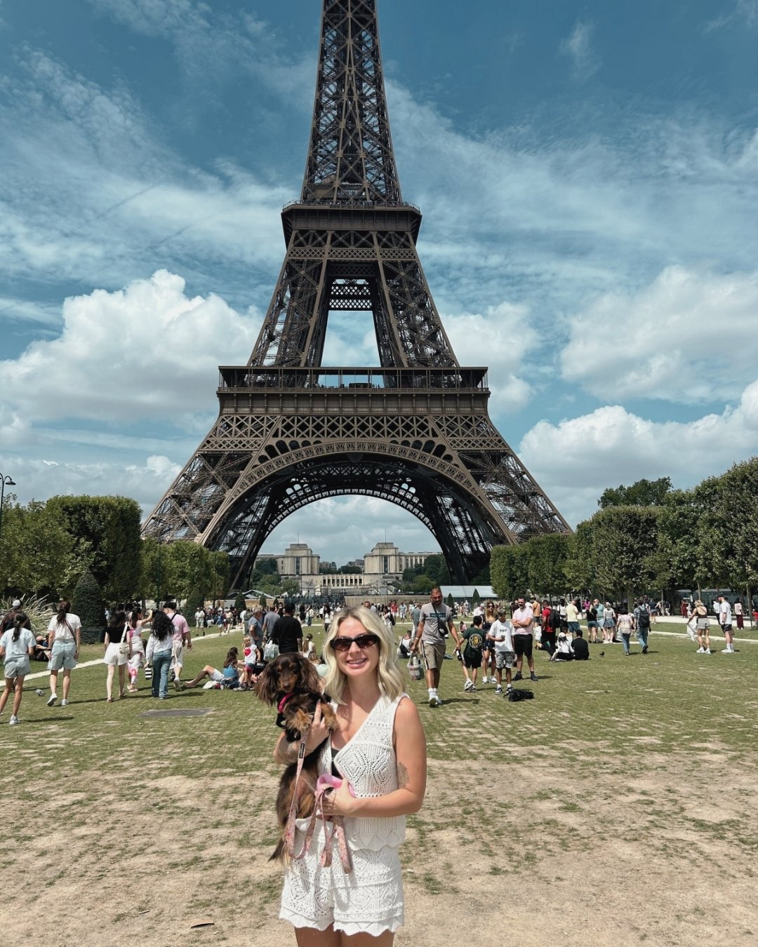 A woman holding a small dog in front of the Eiffel Tower in Paris, France, with a crowd of tourists and trees around.