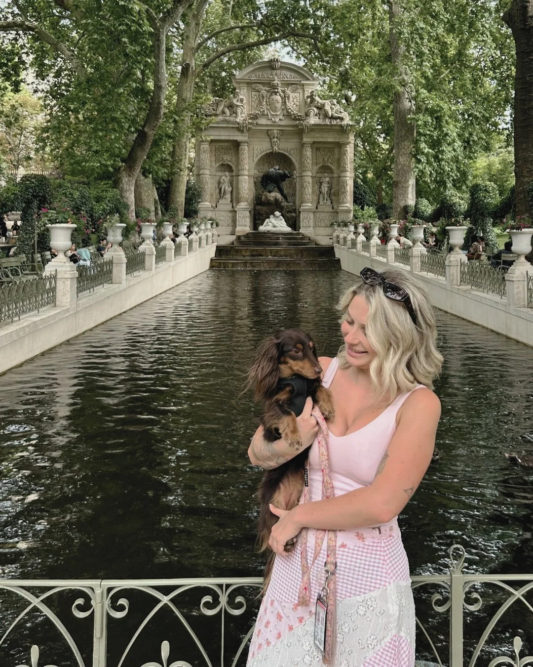 A woman smiling while holding a small brown Dachshund puppy in a park with a decorative fountain and trees in the background.