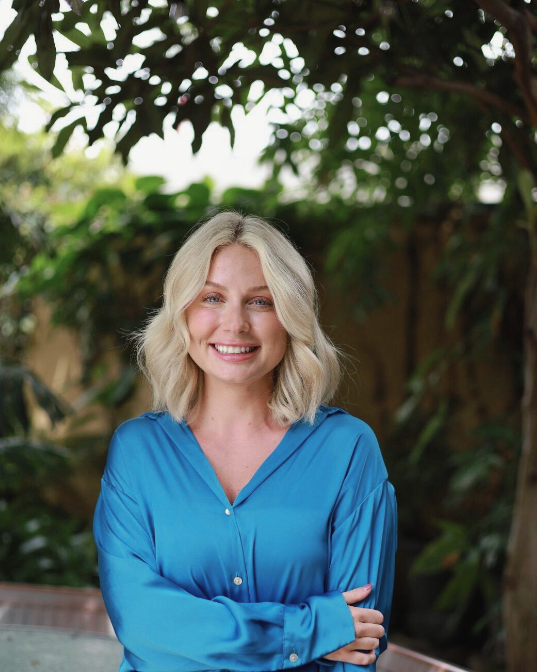 A woman with blonde wavy hair wearing a bright blue satin blouse, smiling with arms crossed, in a lush garden setting with green foliage.