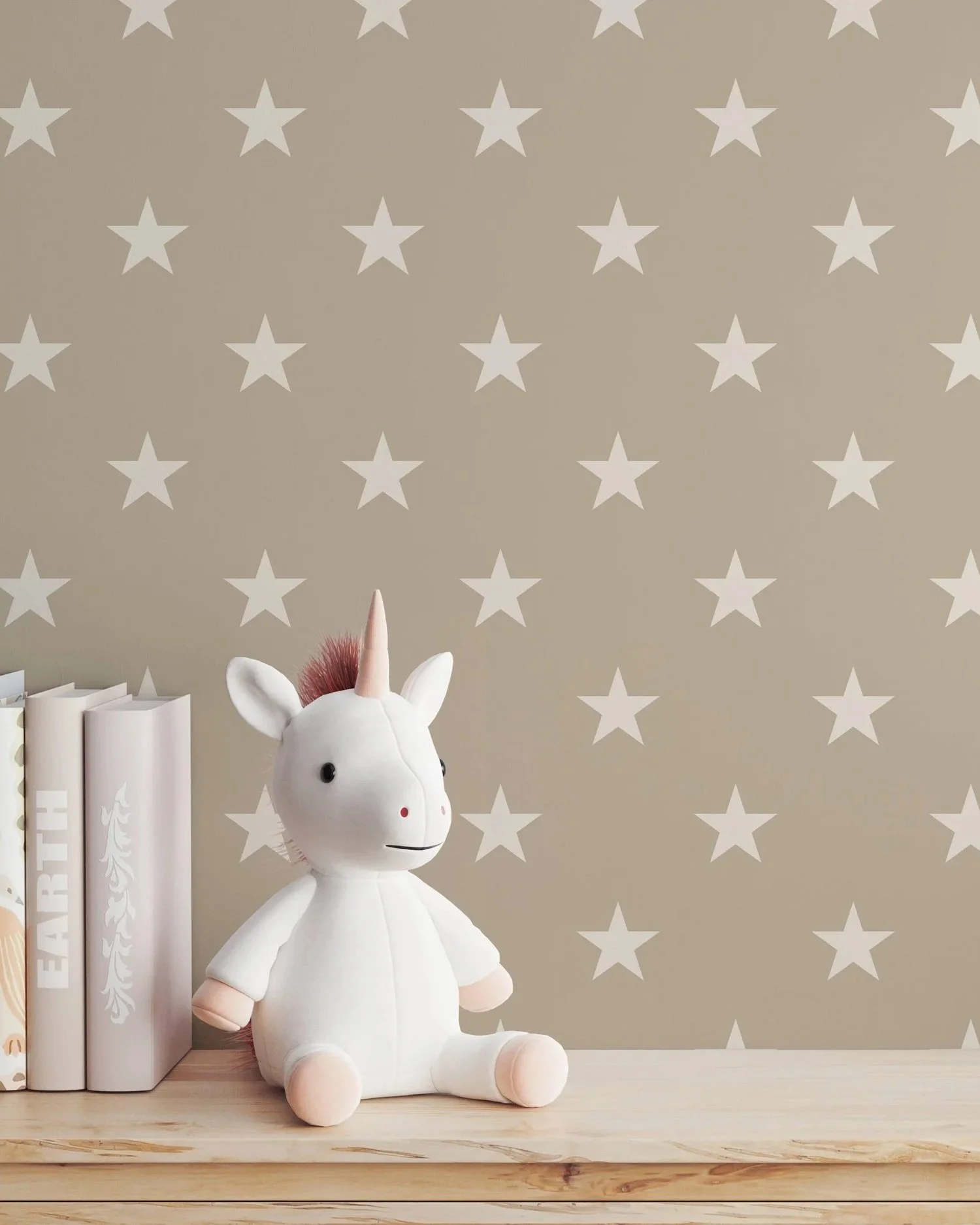 A plush unicorn toy with a white body, pink hair, and a beige horn, sitting on a wooden surface next to three white books with decorative spine designs, against a beige wall with a white star pattern.