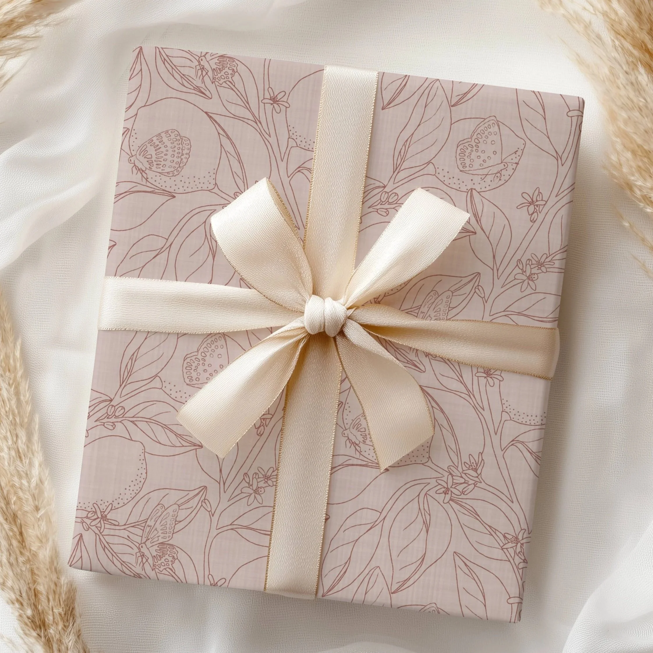 A wrapped gift box with pink floral and butterfly patterns, tied with a cream-colored satin ribbon, placed on a white fabric surface with cream-colored fluffy border.