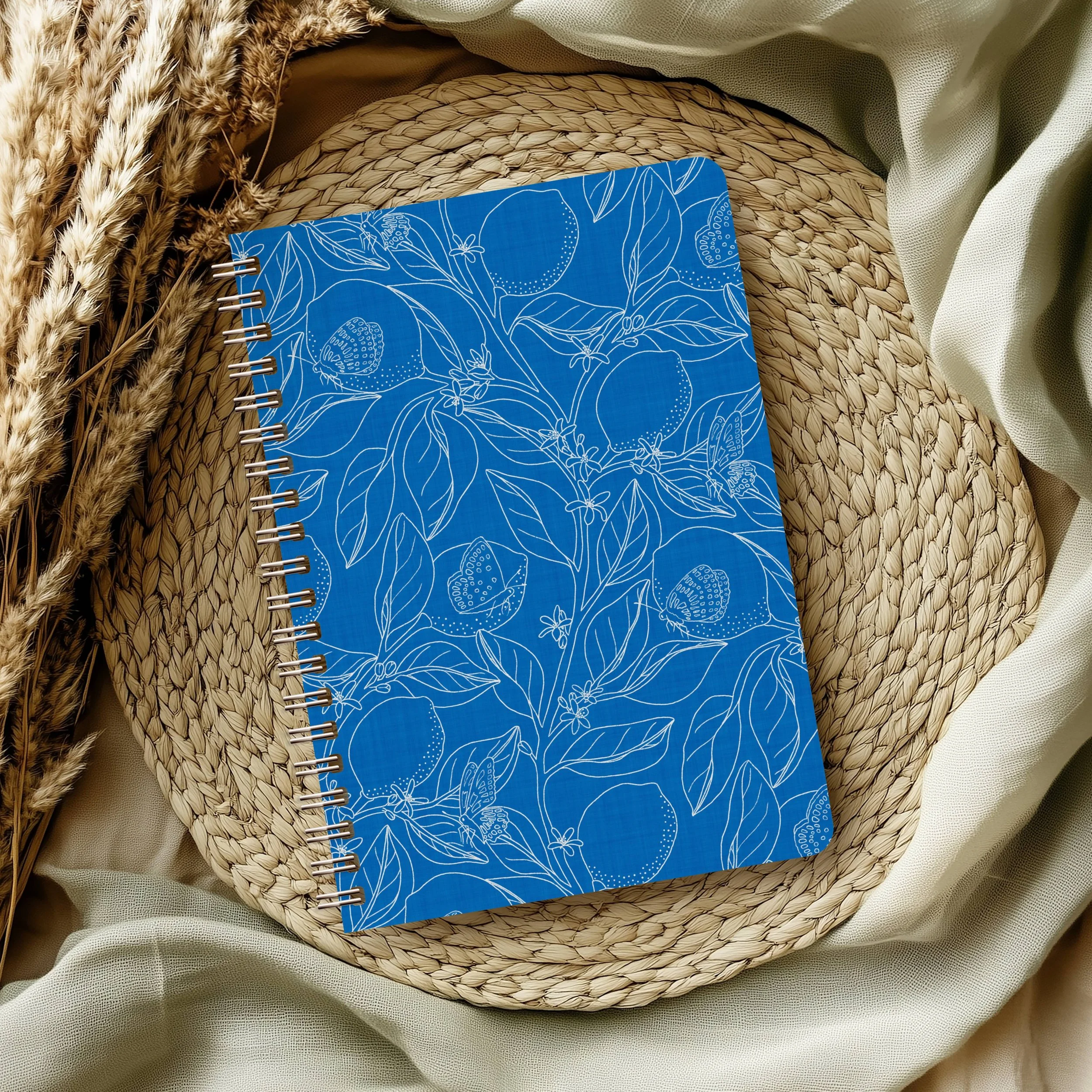 A blue spiral-bound notebook with white botanical line art cover featuring leaves and berries, placed on top of a woven straw placemat, with dried wheat stalks and beige fabric surrounding it.