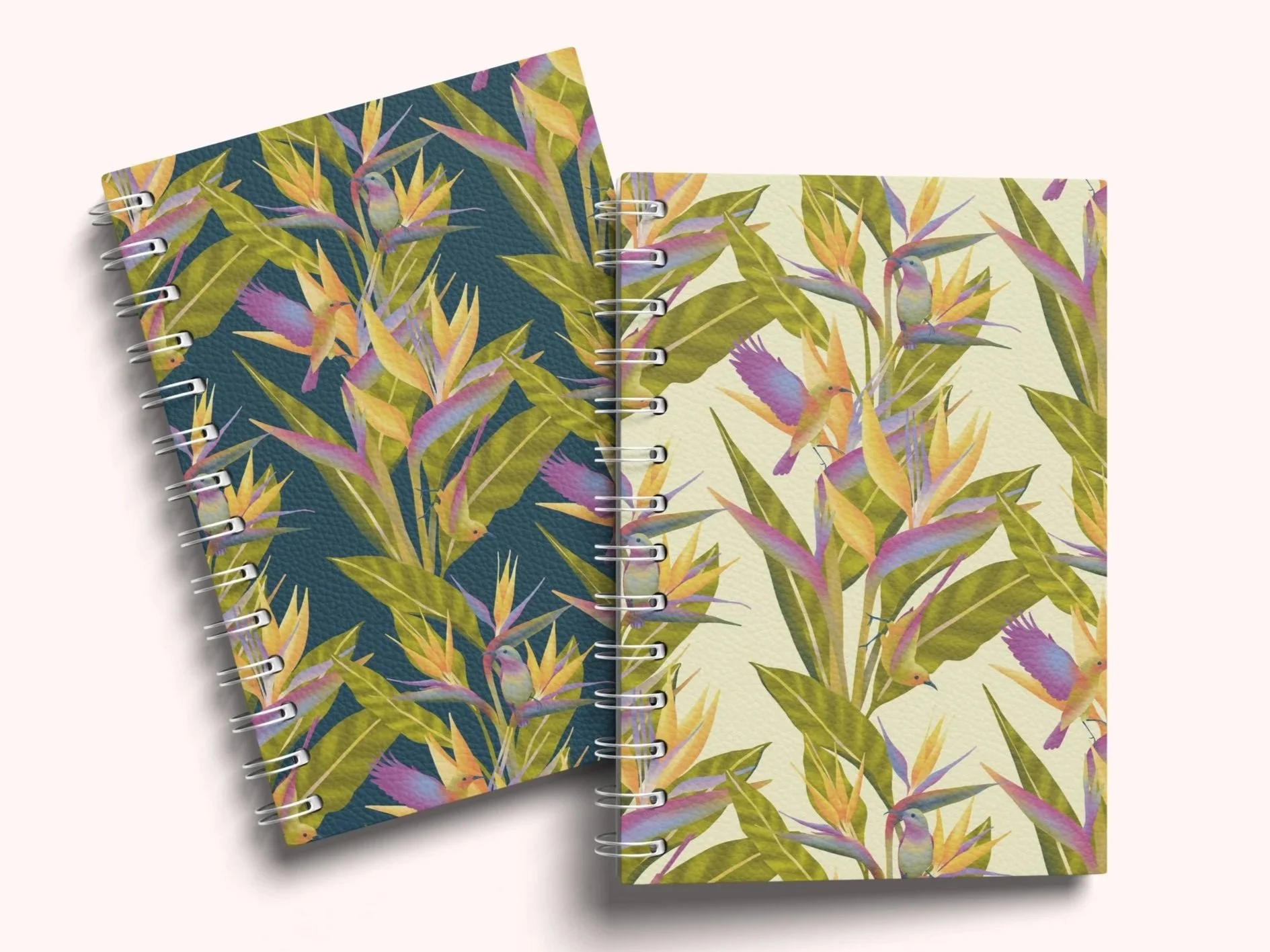 Two spiral-bound notebooks with tropical bird and plant designs, one with a navy blue background and the other with a light yellow background, placed on a white surface.