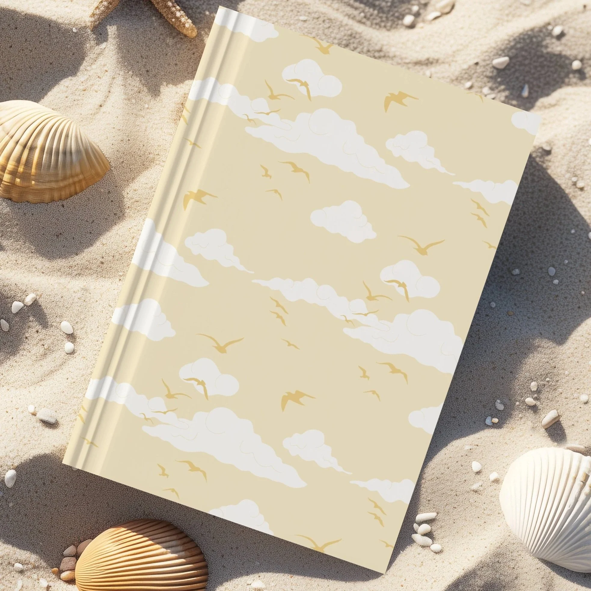 A notebook with a cover featuring a pattern of white clouds and golden birds, lying on sandy beach surrounded by seashells and small pebbles.