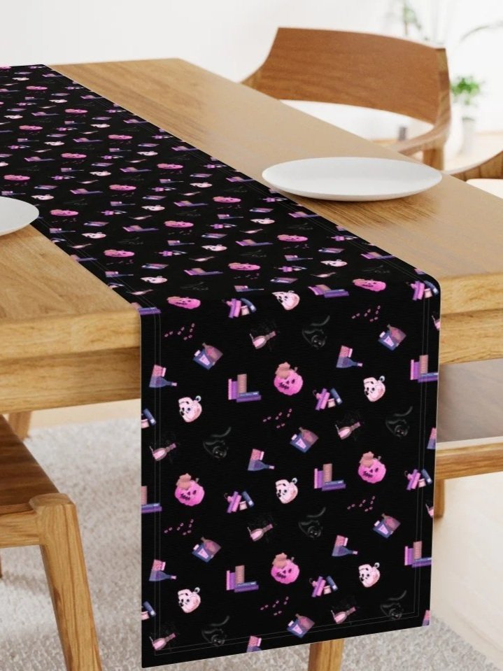 A black table runner with Halloween-themed images including pink pumpkins, white ghosts, purple candles, and black bats, runs down the center of a wooden dining table with white plates.