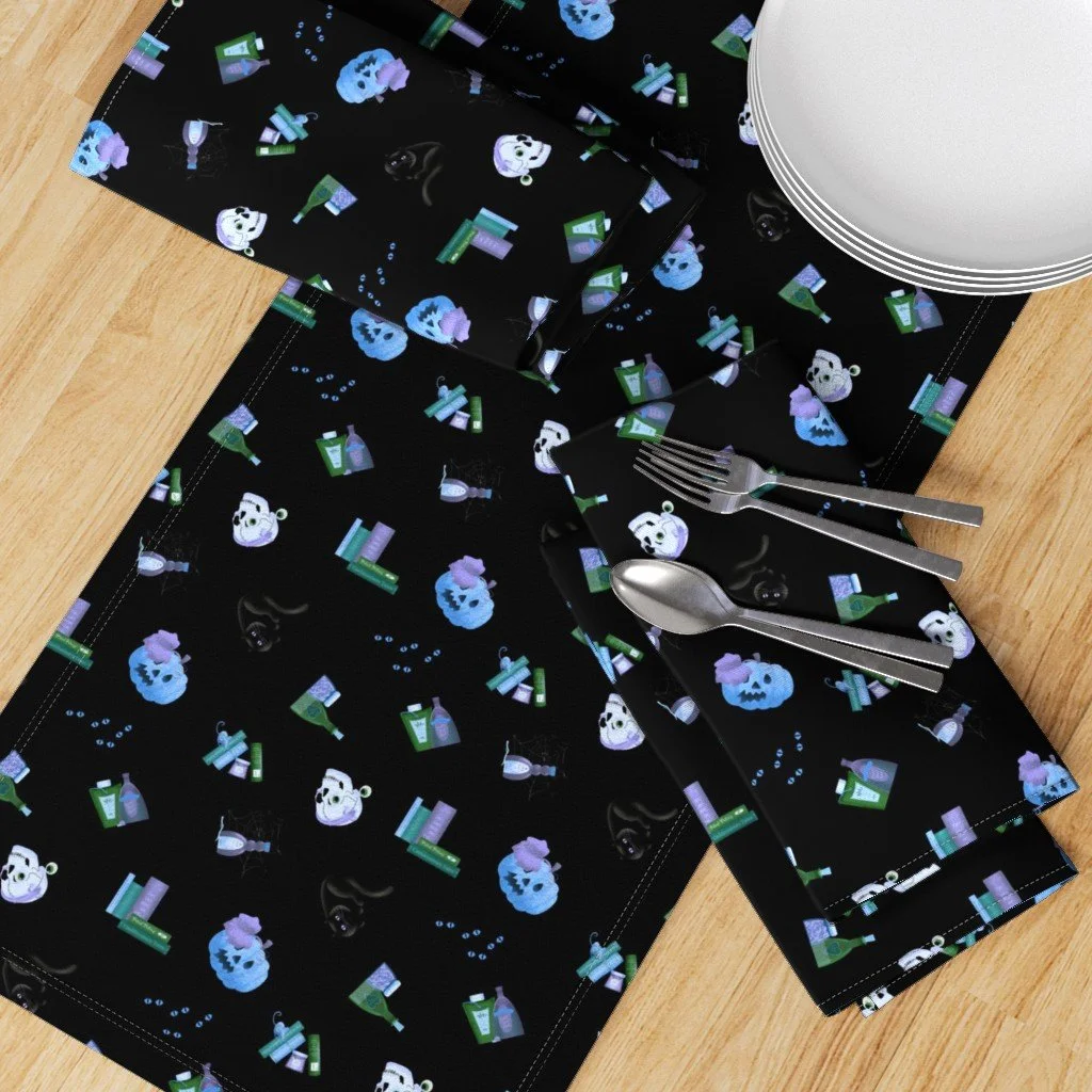 Halloween-themed table setting with black paper placemats decorated with blue and purple pumpkin characters, medicine bottles, and bubbling potion images. The table includes a white plate, a fork, and a spoon on the placemats, with a stack of papers or napkins underneath.