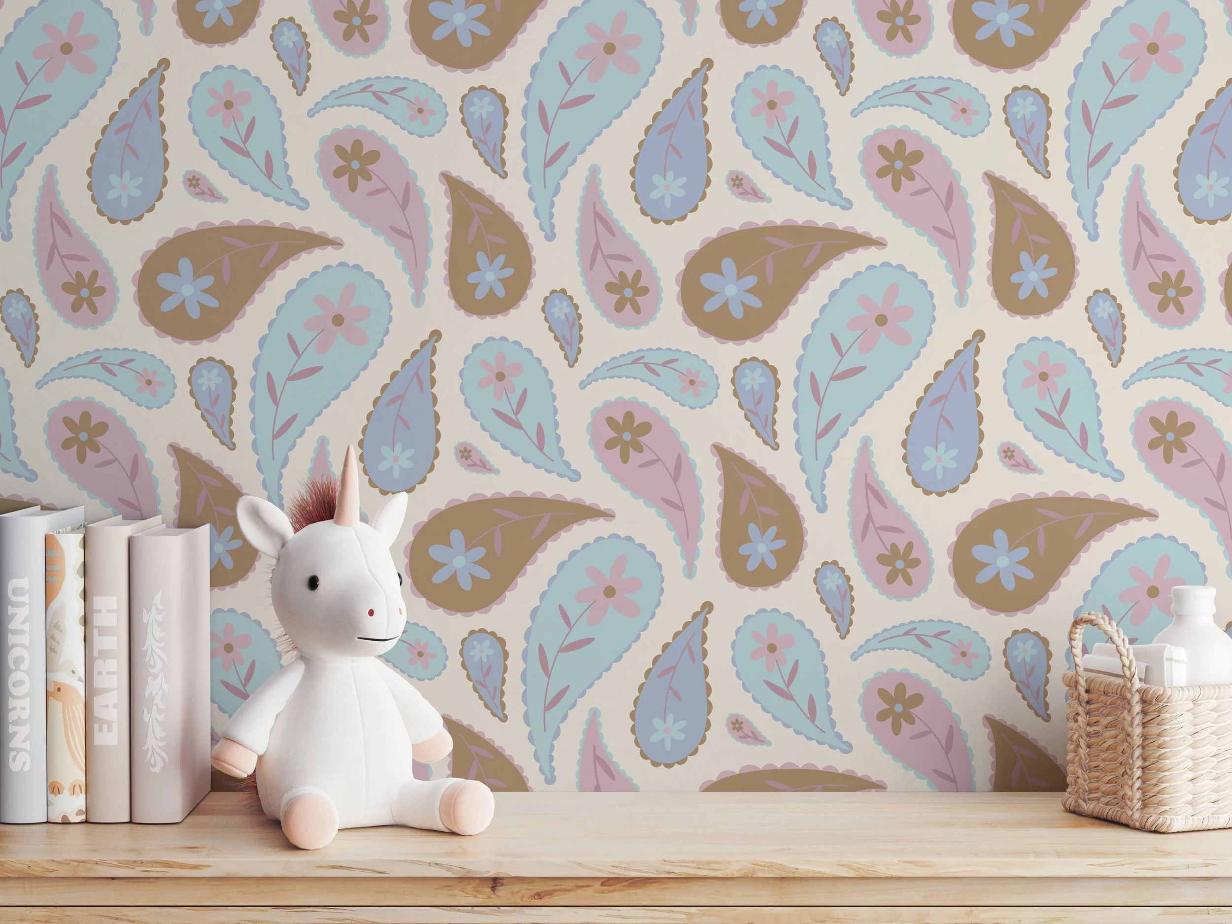 A plush unicorn toy with a white body, pink mane, and a golden horn sits on a wooden surface next to white books and a woven basket, with a pastel paisley patterned wallpaper in the background.