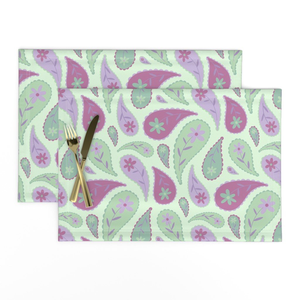 Two pastel-colored placemats with a purple and green floral paisley pattern, set with a fork and knife crossed on top.