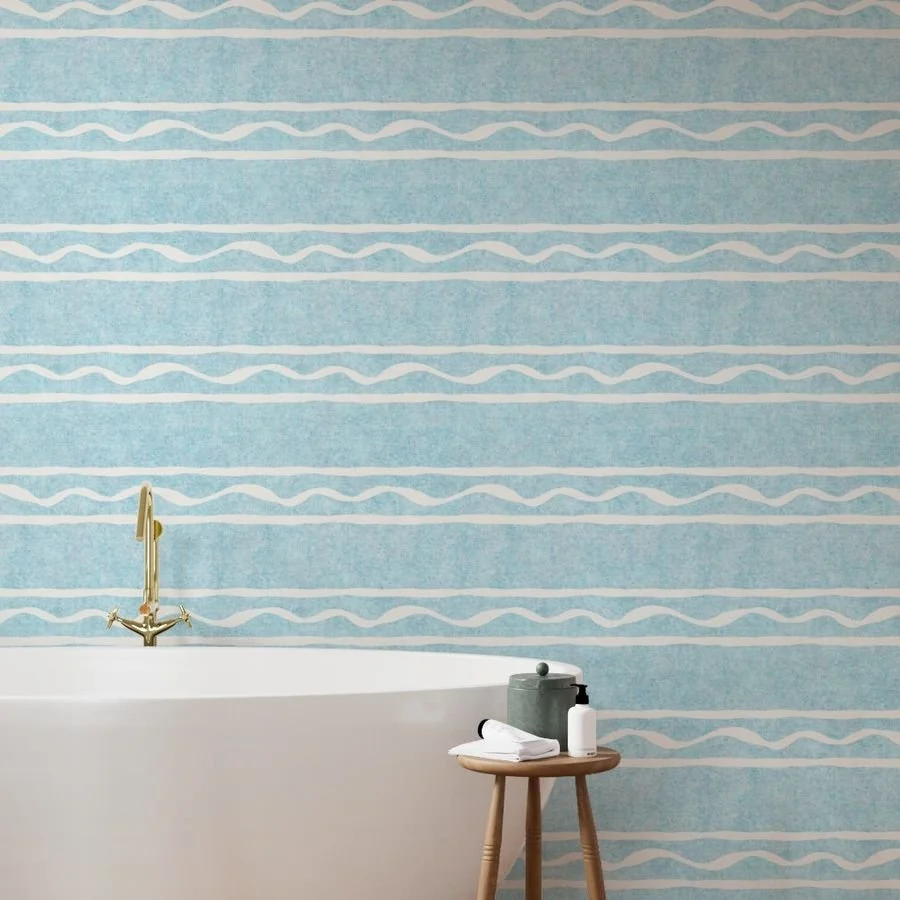 A bathroom with a white bathtub, a gold faucet, and a small wooden side table holding soap and towels. The wall has blue wallpaper with white wavy lines.