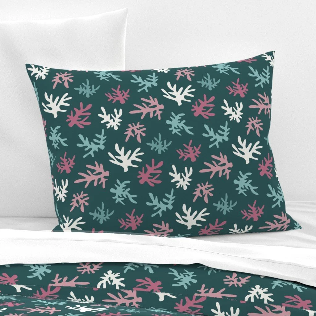 Decorative pillow with a dark teal background and a pattern of pink, white, and light blue abstract leaves, placed on a bed with white sheets and pillows.