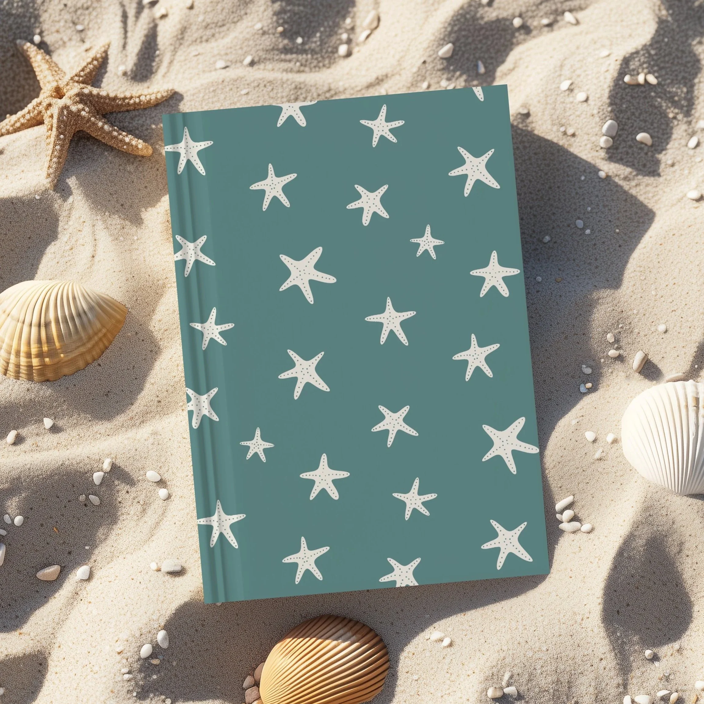 A notebook with a starfish pattern on the cover, placed on sand alongside seashells and small shells, with parts of a person's foot visible at the bottom.