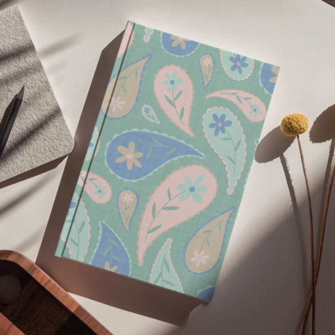 A pastel-colored notebook with a floral, paisley pattern on the cover, placed on a white surface with dried flowers nearby.