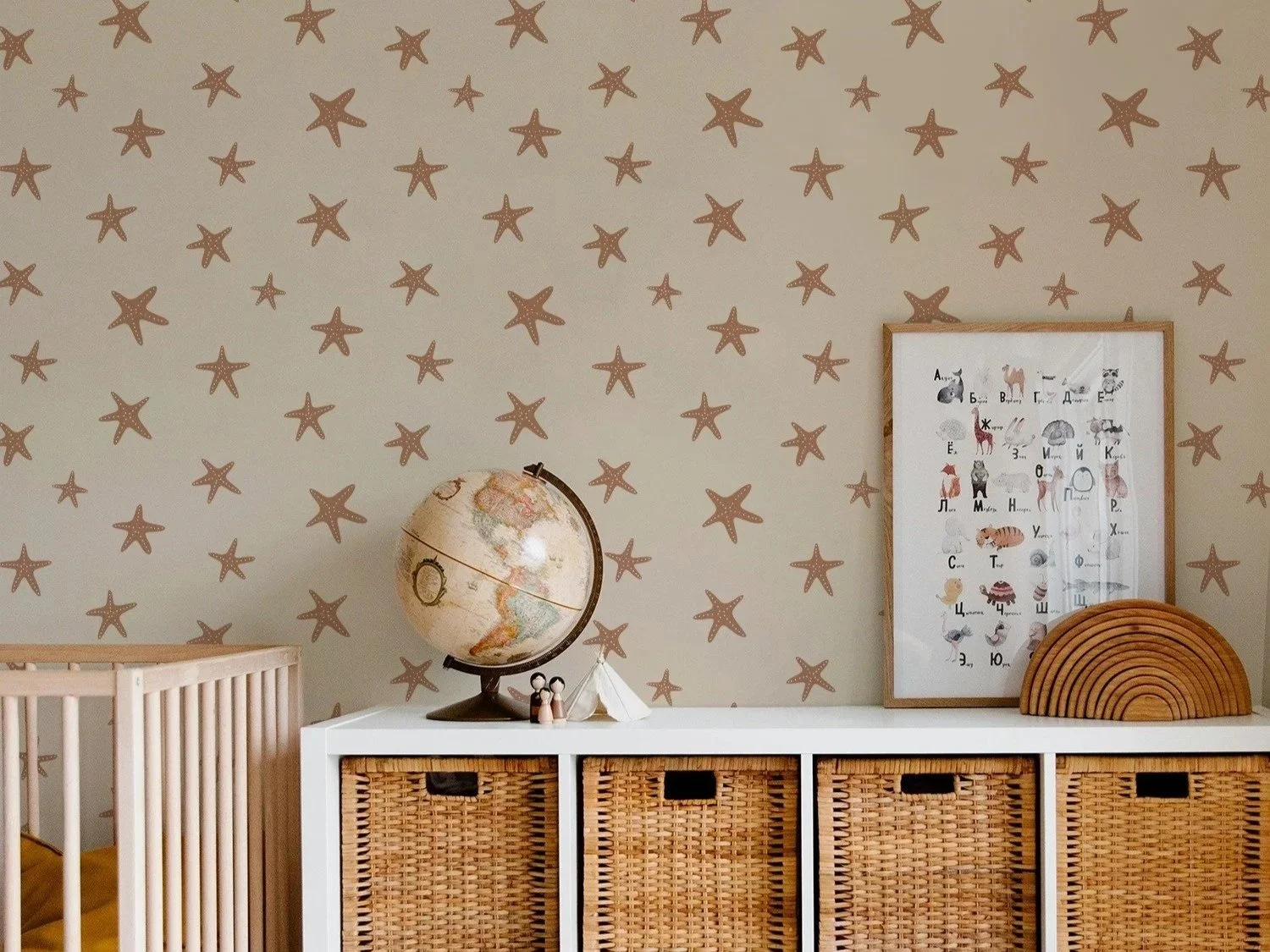 A nursery wall decorated with starfish wallpaper, a globe, a framed animal alphabet poster, a fan-shaped wooden decoration, and a white storage unit with wicker baskets.
