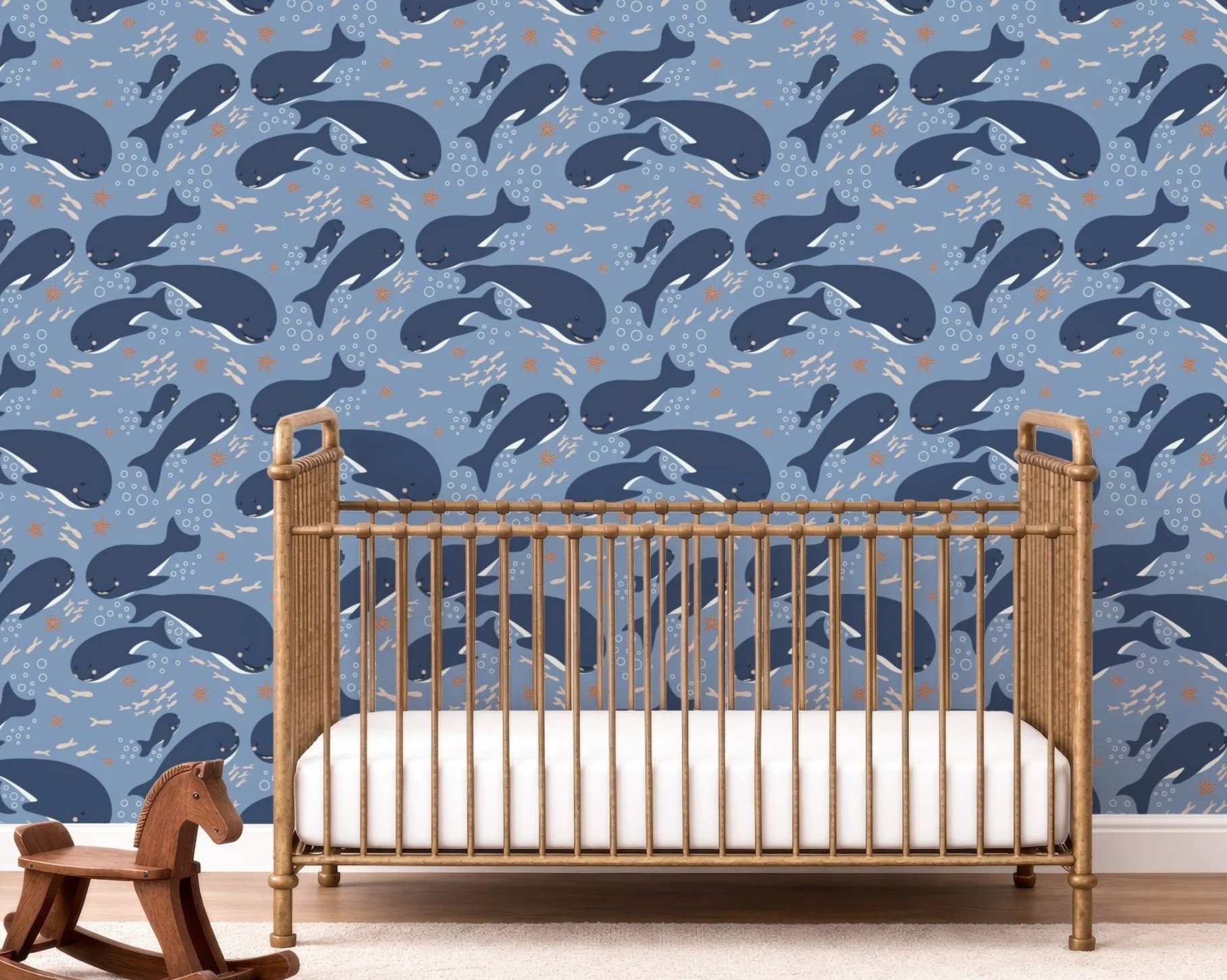 A nursery with a blue whale-themed wallpaper featuring various sizes of whales, fish, and starfish. In the foreground, there is a wooden crib with a white mattress, and to the left, there is a wooden rocking horse with a carved horse head design on a beige carpeted floor.