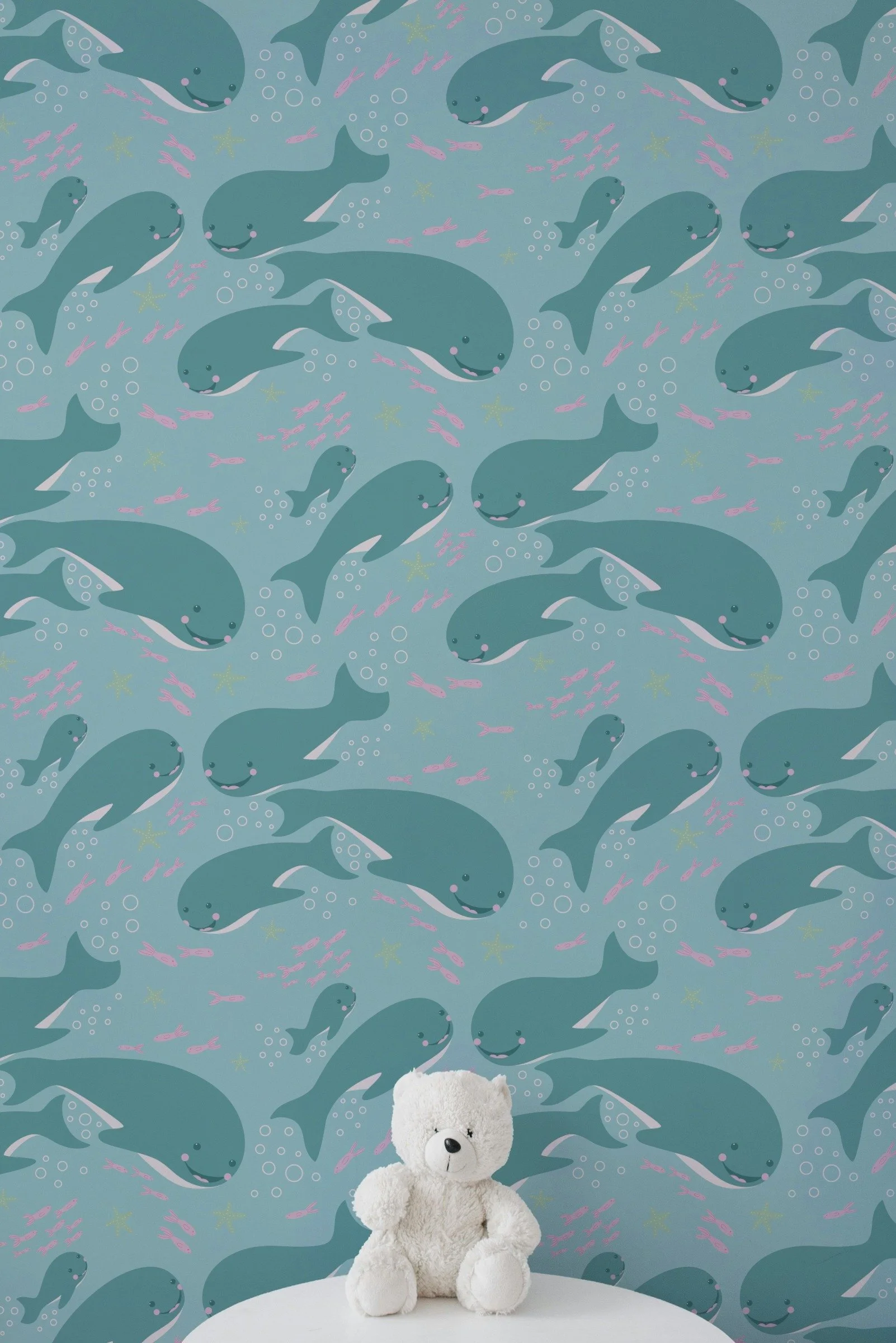 A white teddy bear sitting on a white surface in front of a blue wallpaper decorated with smiling whales, pink fish, bubbles, and starfish.
