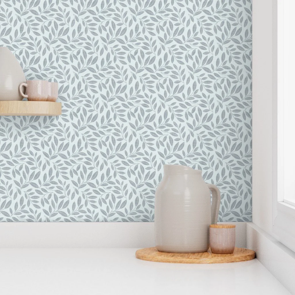 A beige pitcher and a small cup on a round wooden tray on a white surface near a window, with a wall covered in light blue and white leaf-patterned wallpaper.