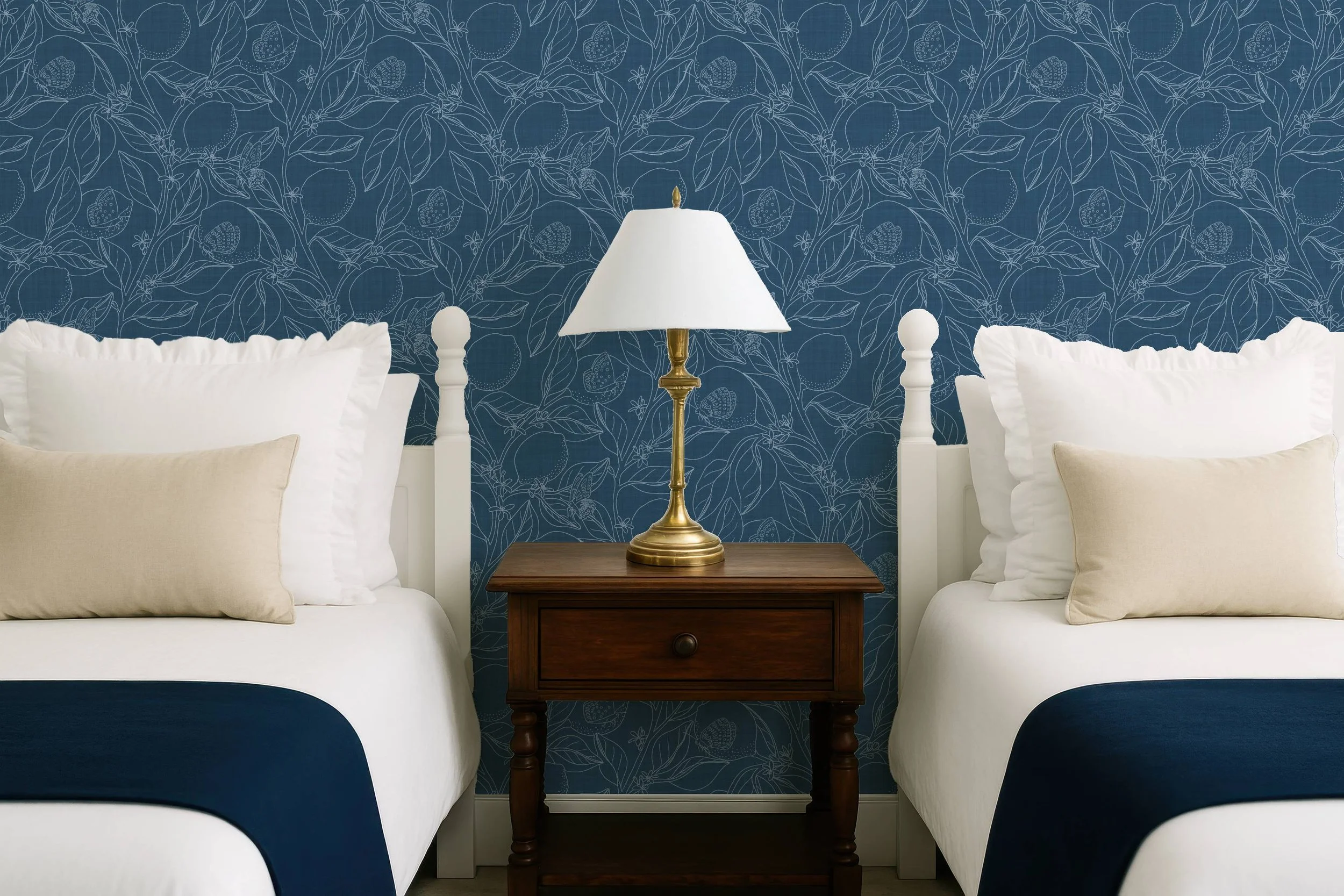 Two beds with white pillows and beige pillows, separated by a wooden nightstand with a brass lamp, against a blue wallpaper with a fruit and leaf pattern.