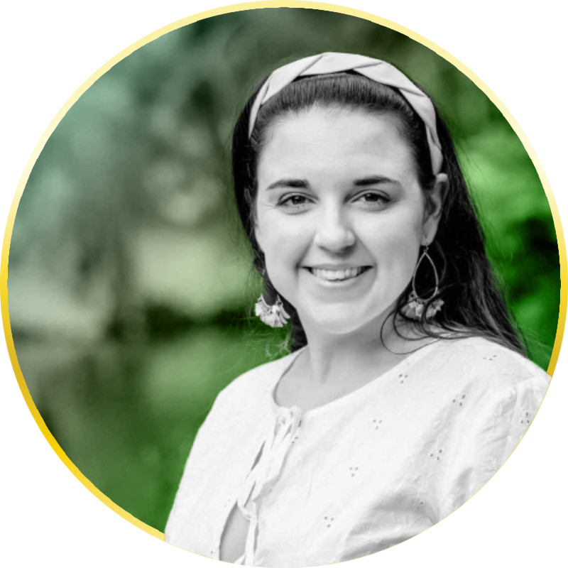 A smiling woman with dark hair pulled back, wearing hoop earrings, a white top, and a headband, outdoors with blurred green foliage in the background.