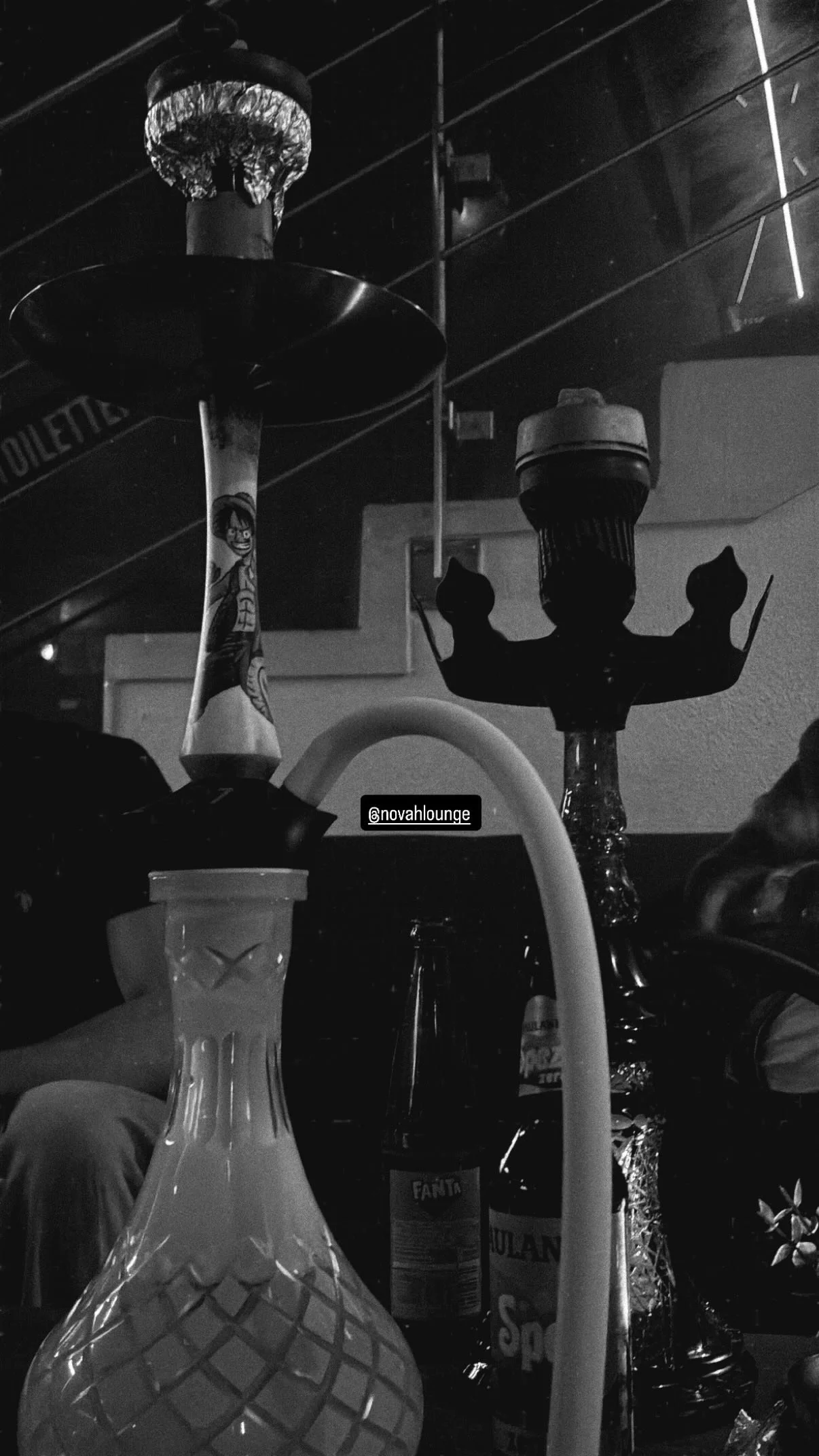 Schwarzer Hintergrund mit Wasserpfeife, Gläsern, Flaschen und Kerzen auf einem Tisch, Stimmung in einer Lounge oder Bar, Instagram-Handle @novahlounge.