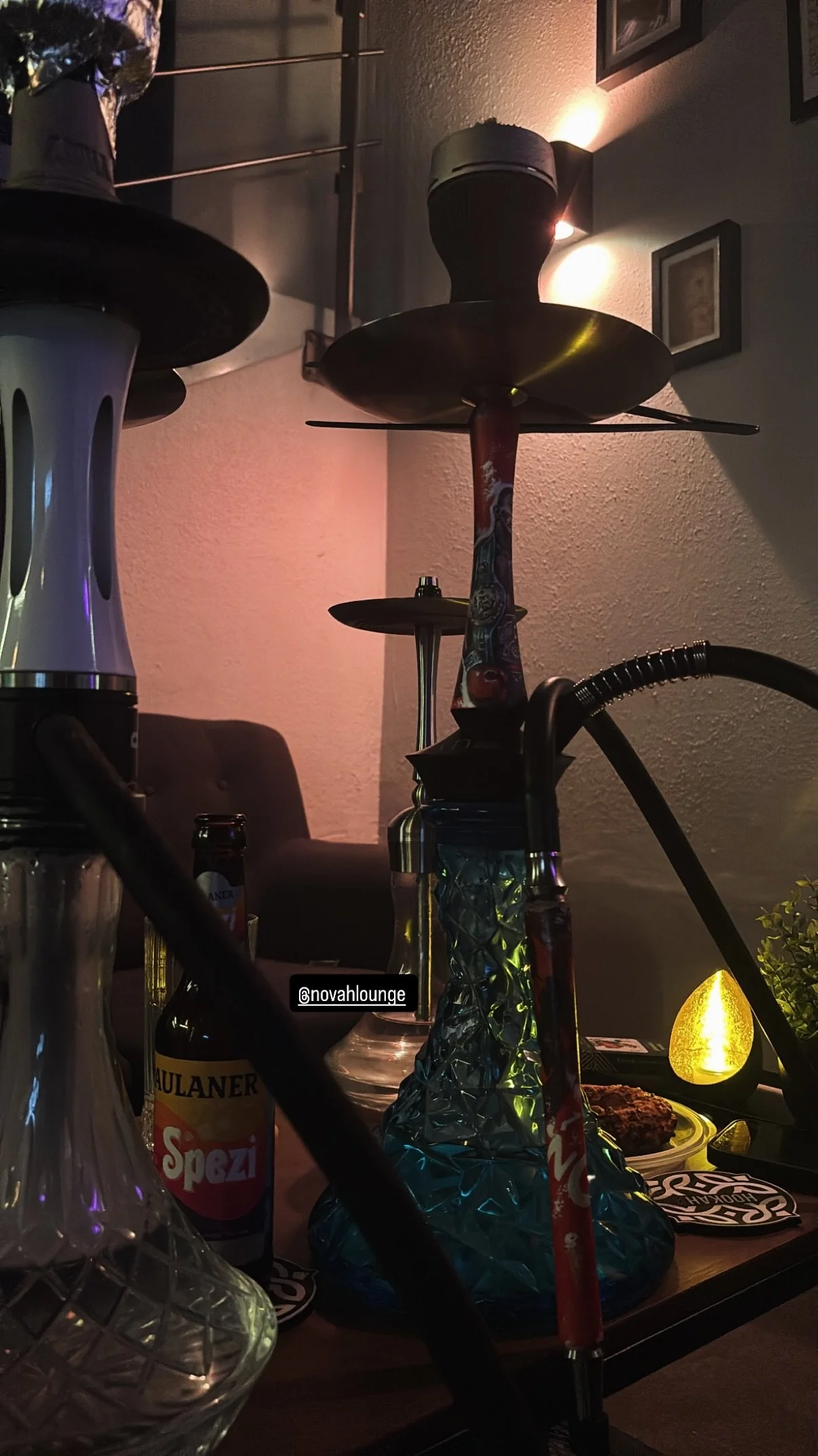 Rauchende Hookah auf einem Tisch, umgeben von Getränken und Snacks in einer dunklen Lounge. Es gibt Kerzenlicht und Bilder an der Wand.