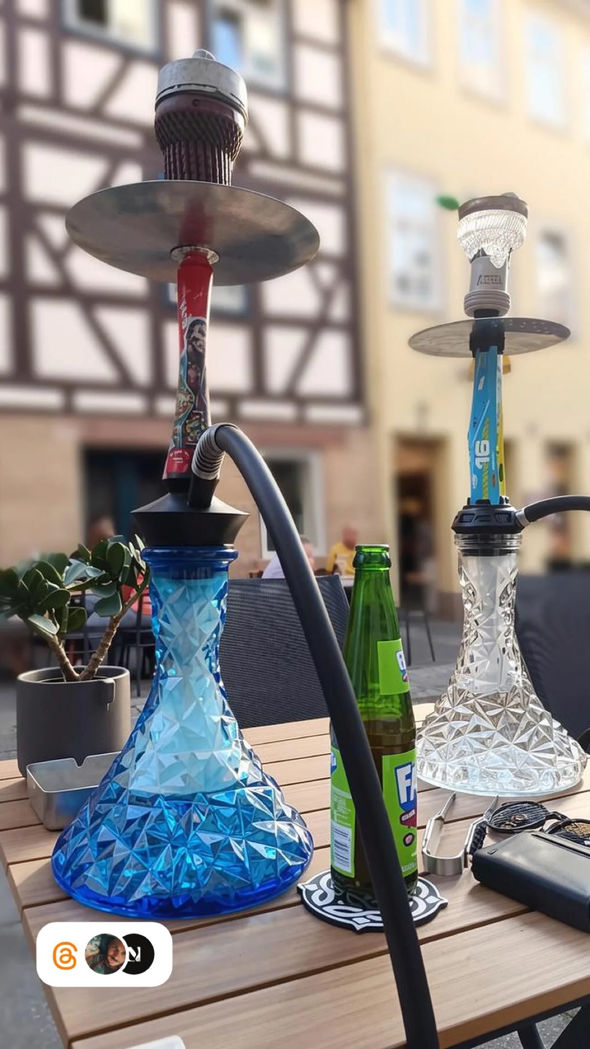 Zwei Hookahs auf einem Holztisch im Freien, begleitet von einer kleinen Pflanze in einem Topf und mehreren Getränkeflaschen, unter hellem Tageslicht vor einem Gebäude.
