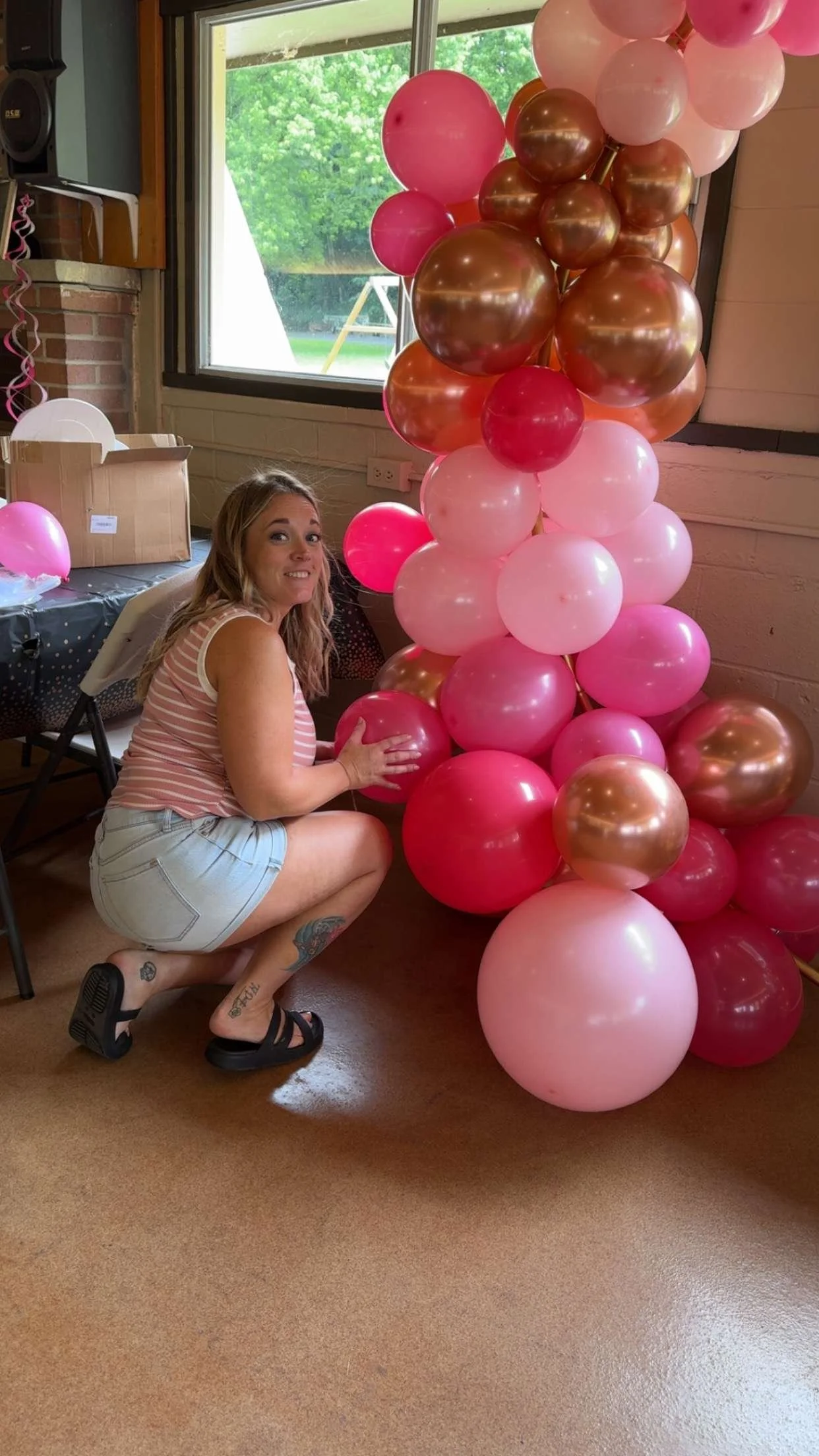 A woman with tattoos and wearing a striped tank top and shorts crouching beside a large arrangement of pink, white, and gold balloons inside a room with a window and a table with more balloons.