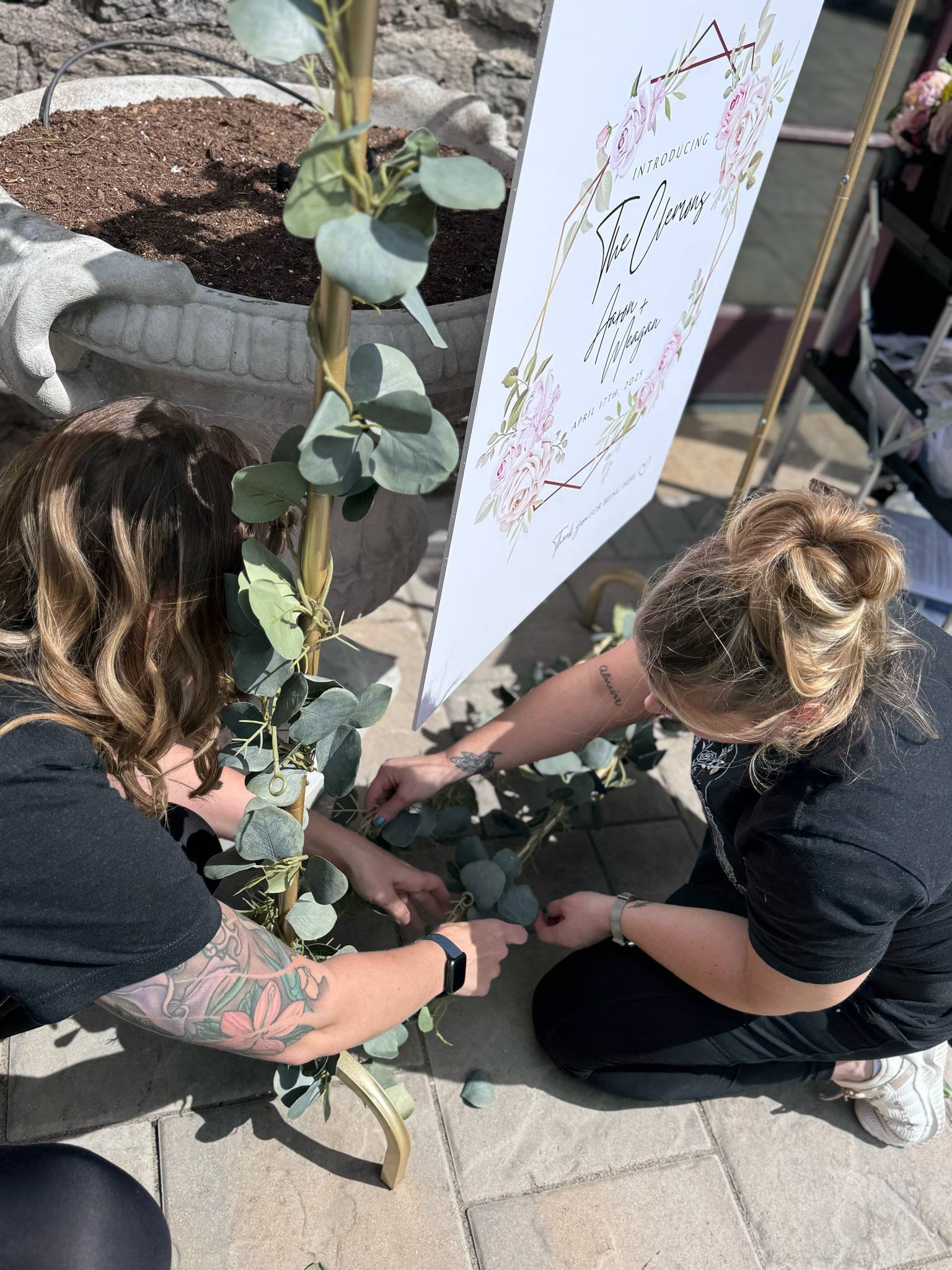 Two women decorate a welcome sign with greenery at an outdoor wedding reception.