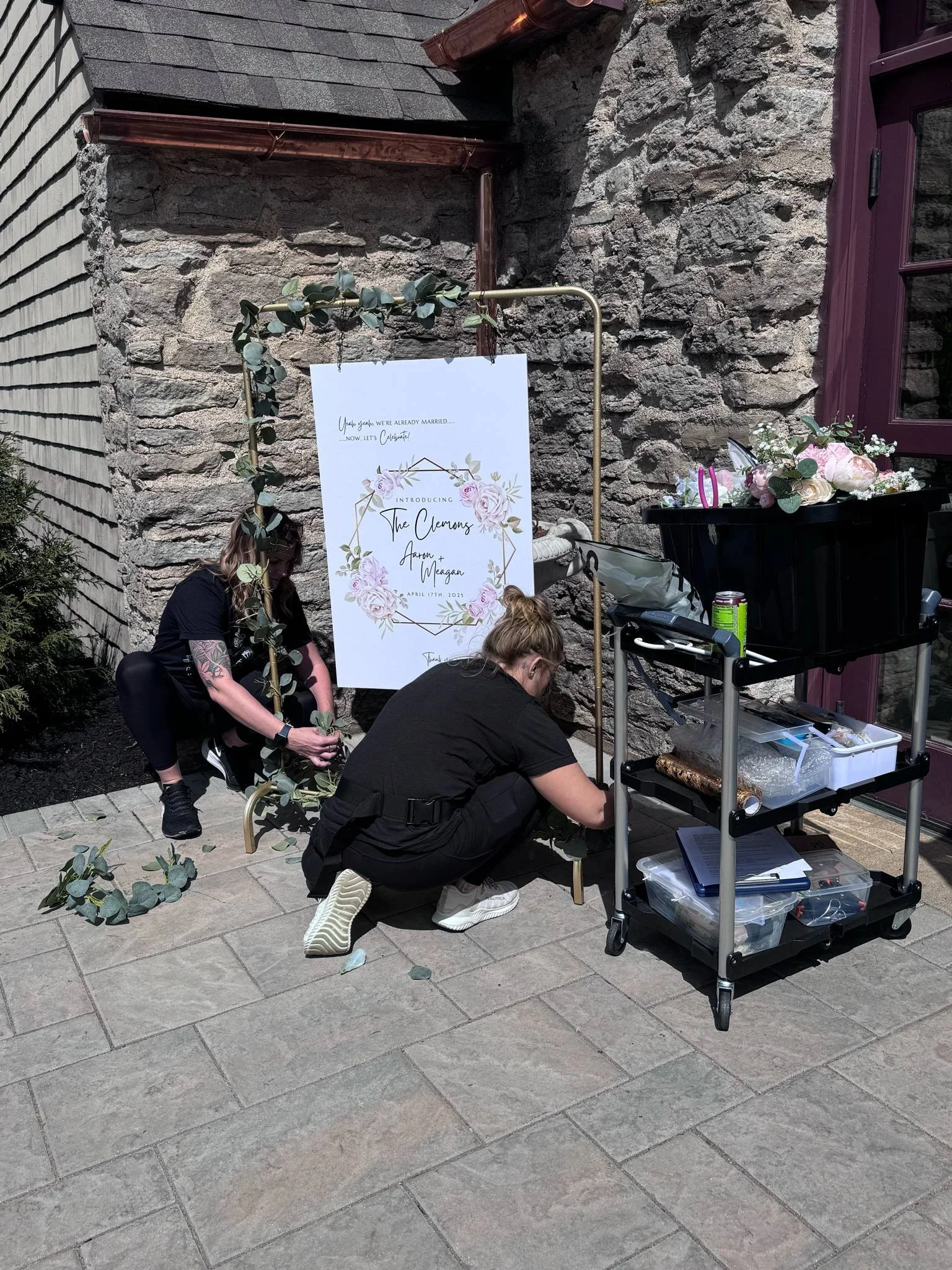 Two women are decorating a floral wedding sign on a brick patio, with one woman crouching and the other sitting, near a cart with flowers and supplies, against a stone wall.
