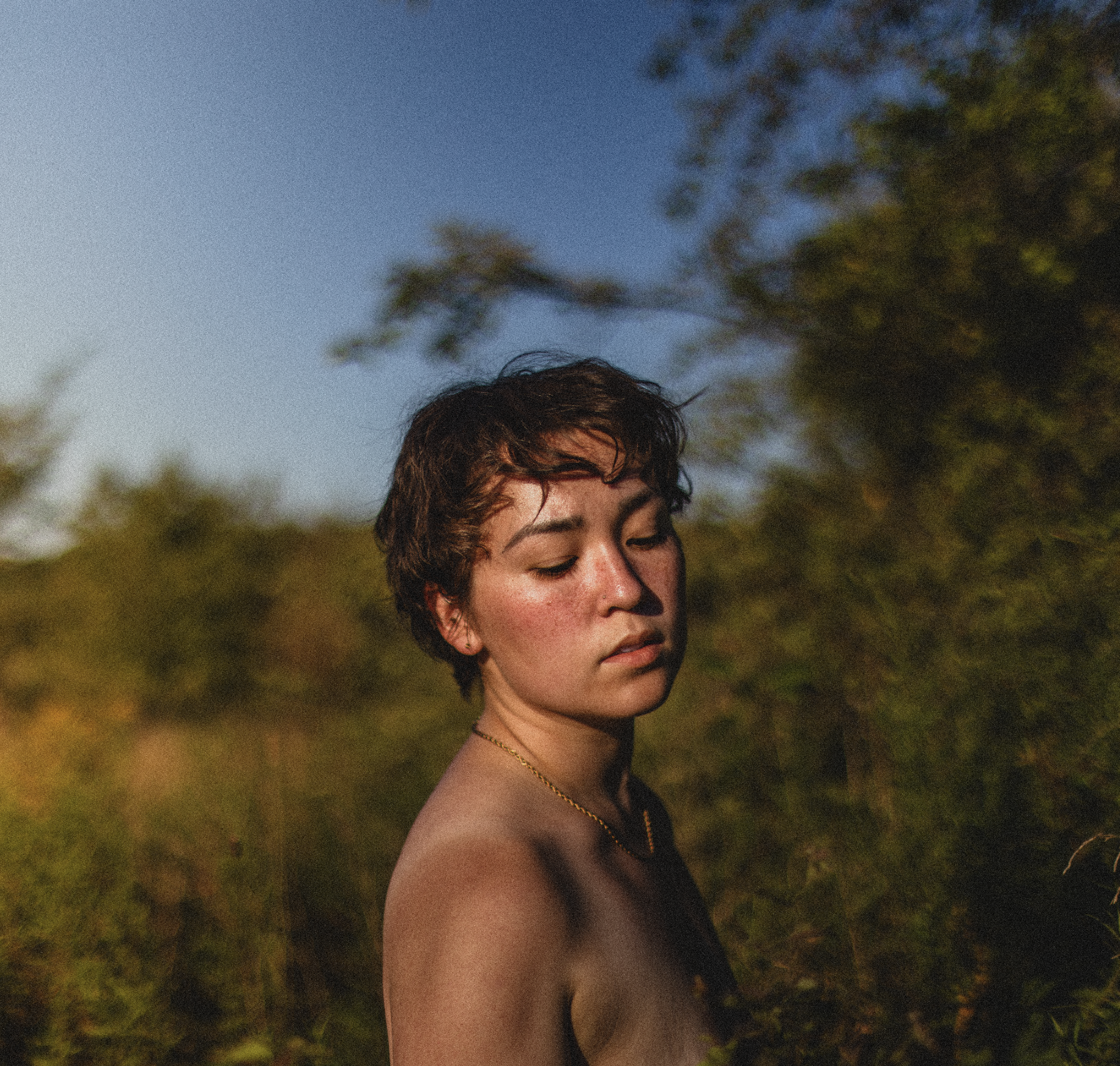 A young femme person with short brown hair and closed eyes standing outdoors among trees with a clear blue sky in the background.