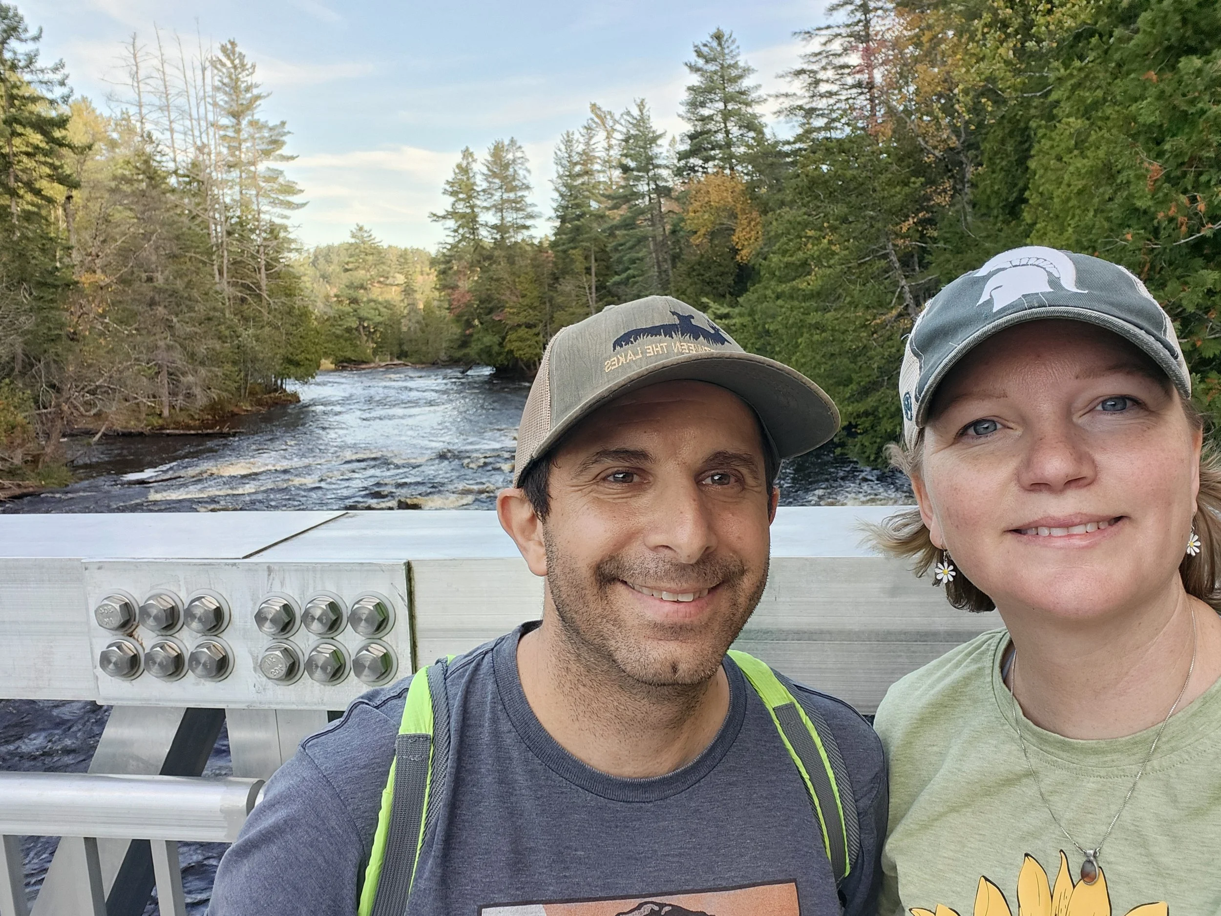 A smiling man and woman taking a selfie on a bridge over a river surrounded by trees with autumn foliage.