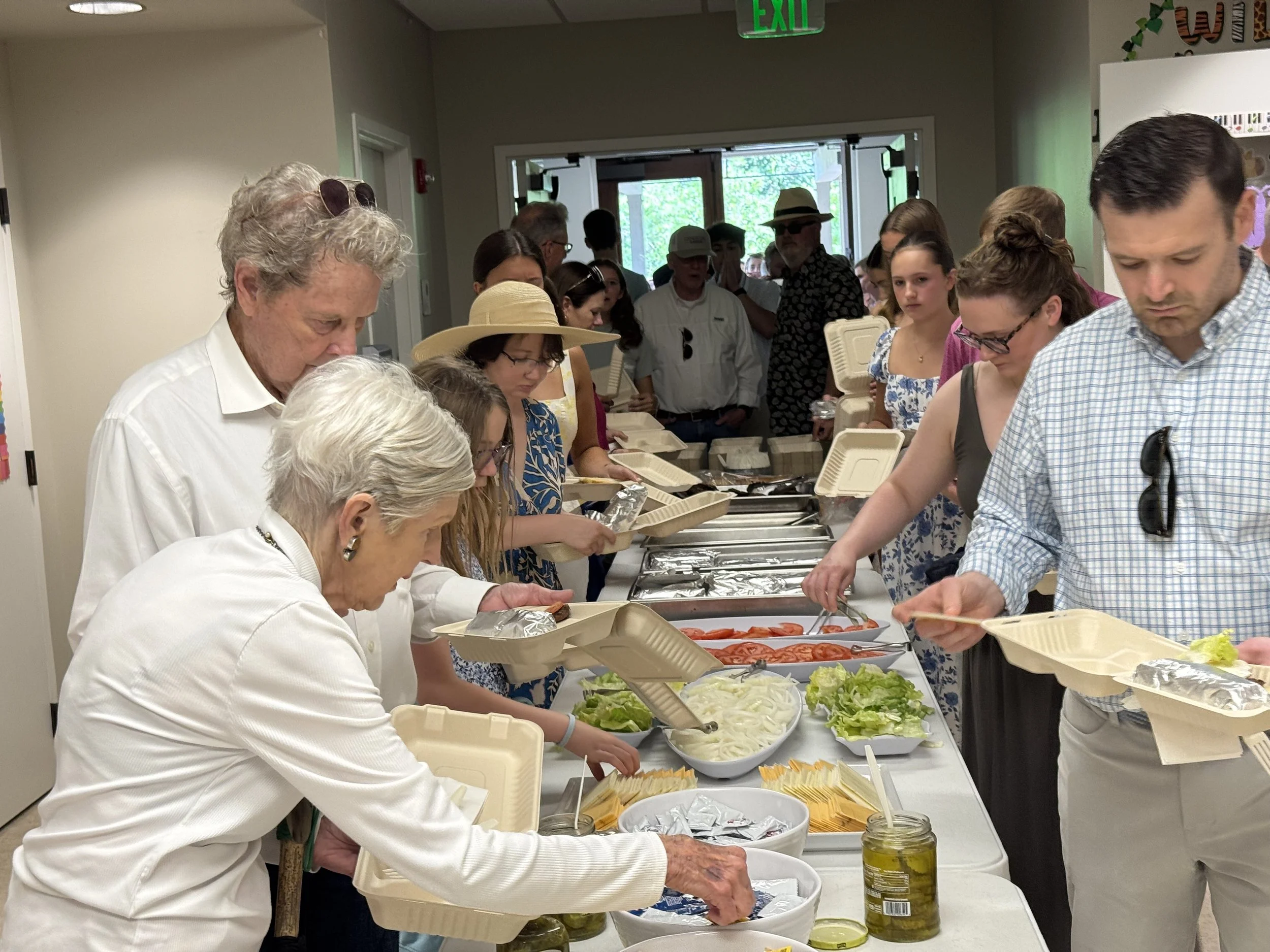 People standing in line getting food from a buffet table at a gathering or party in a well-lit indoor space.