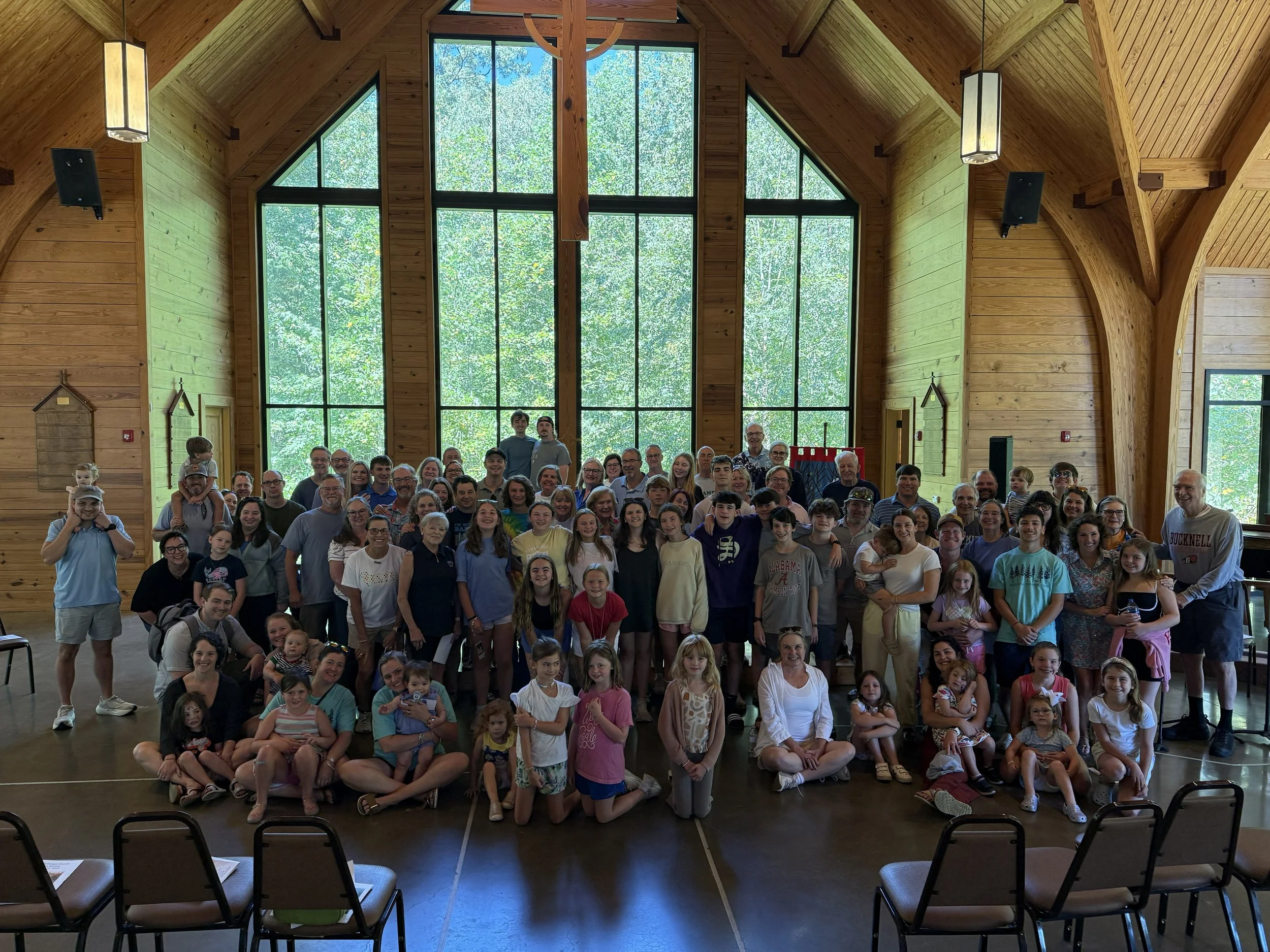 Large group of people of various ages gathered inside a wooden hall with tall windows, posing for a group photo.