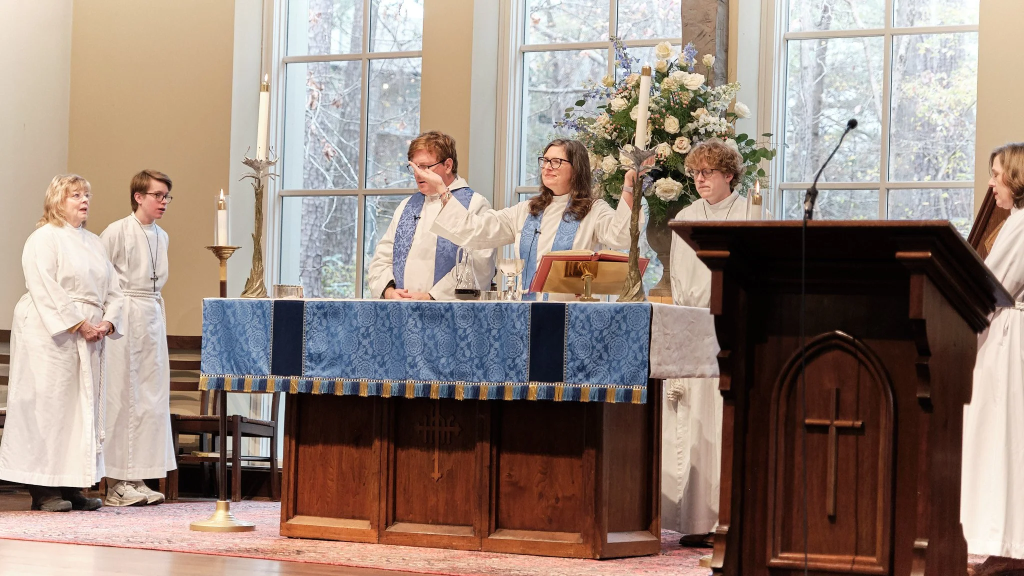 A religious ceremony, likely a Christian church service, with a priest and altar servers gathered around an altar decorated with a blue and gold cloth, candles, and flowers.