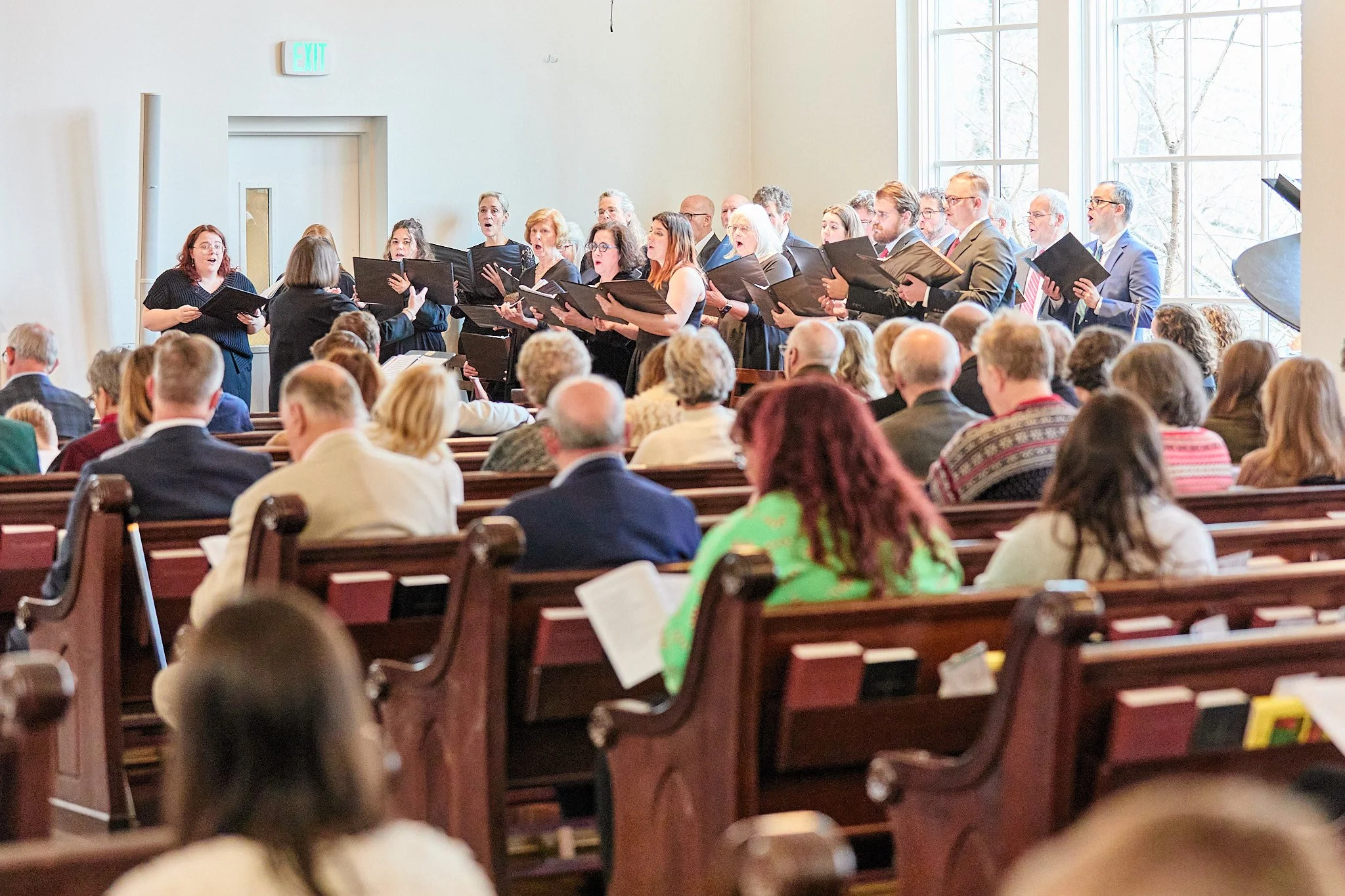 A choir performing inside a church with an audience seated in wooden pews, large windows behind the choir, and an exit sign on the wall.