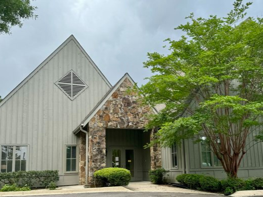 A building with a combination of gray siding and stone exterior, surrounded by green shrubbery and a large tree.