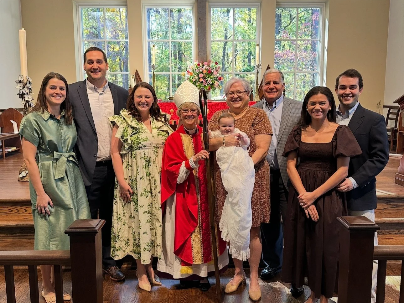 A group of ten people, including a baby, gathered inside a church with large windows and wooden flooring. They are standing in front of an altar decorated with flowers, and the background shows trees outside the windows.