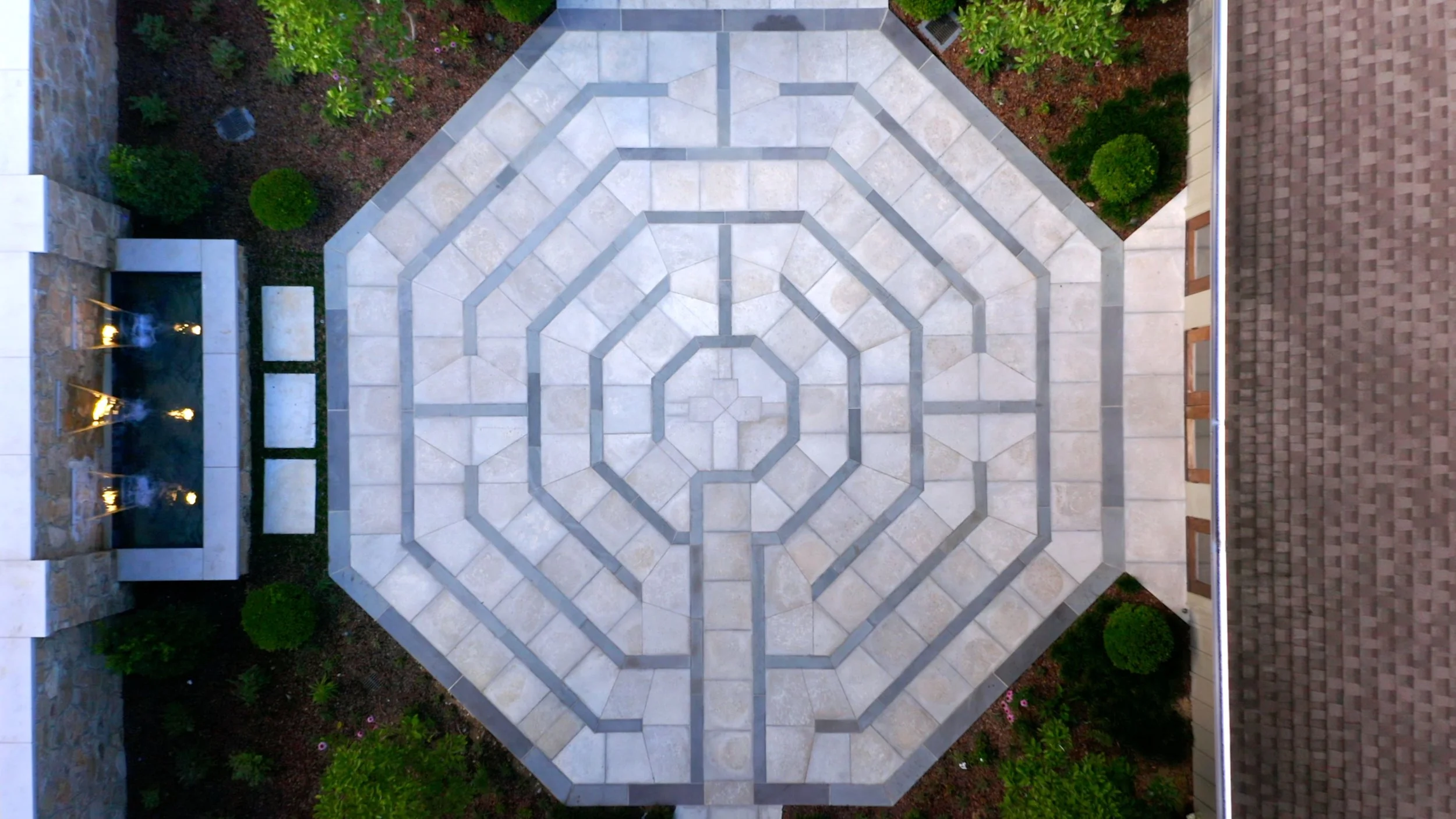 From above view of octagonal tiled patio with a geometric pattern, surrounded by landscaping, a water fountain, and brick and stone building walls.