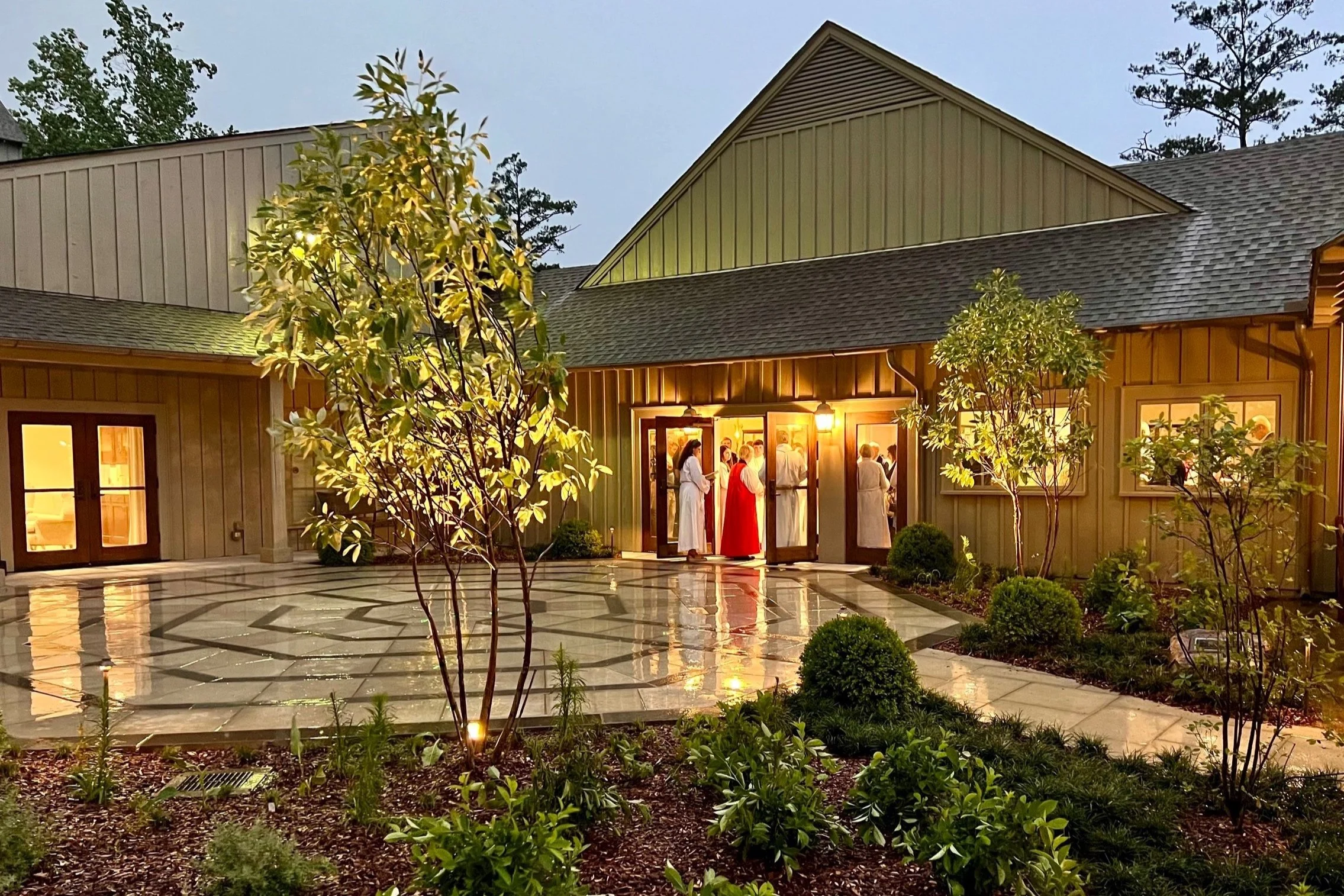 People dressed in white robes and one in a red robe entering a well-lit building with a courtyard, surrounded by plants, trees, and a tiled pathway.