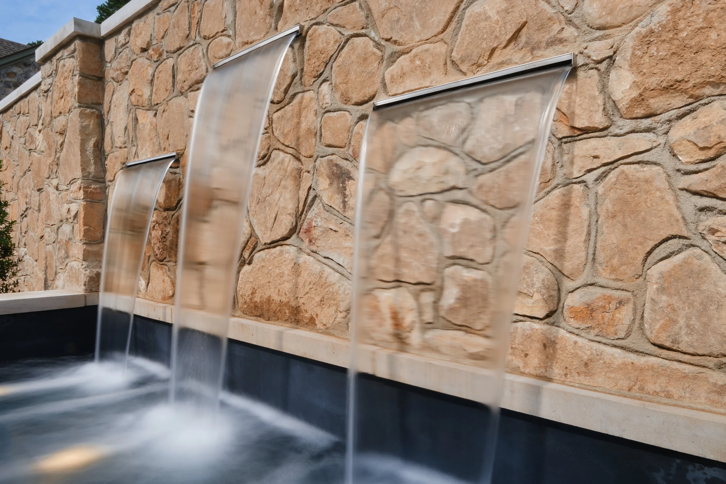 Water features with three vertical streams of water flowing from brushed metal spouts onto a water basin, set against a textured stone wall.