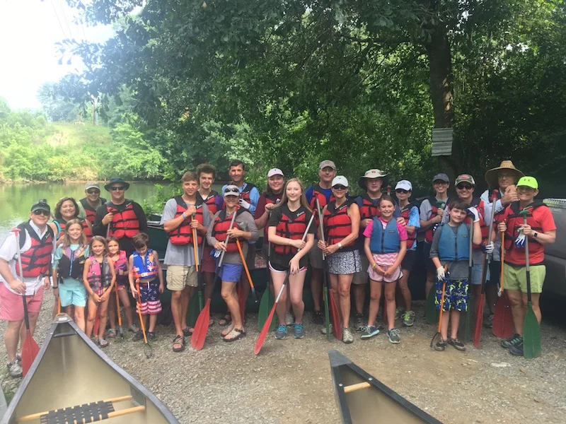 Group of children and adults in life jackets and holding paddles, preparing for a canoeing trip by the river with trees in the background.